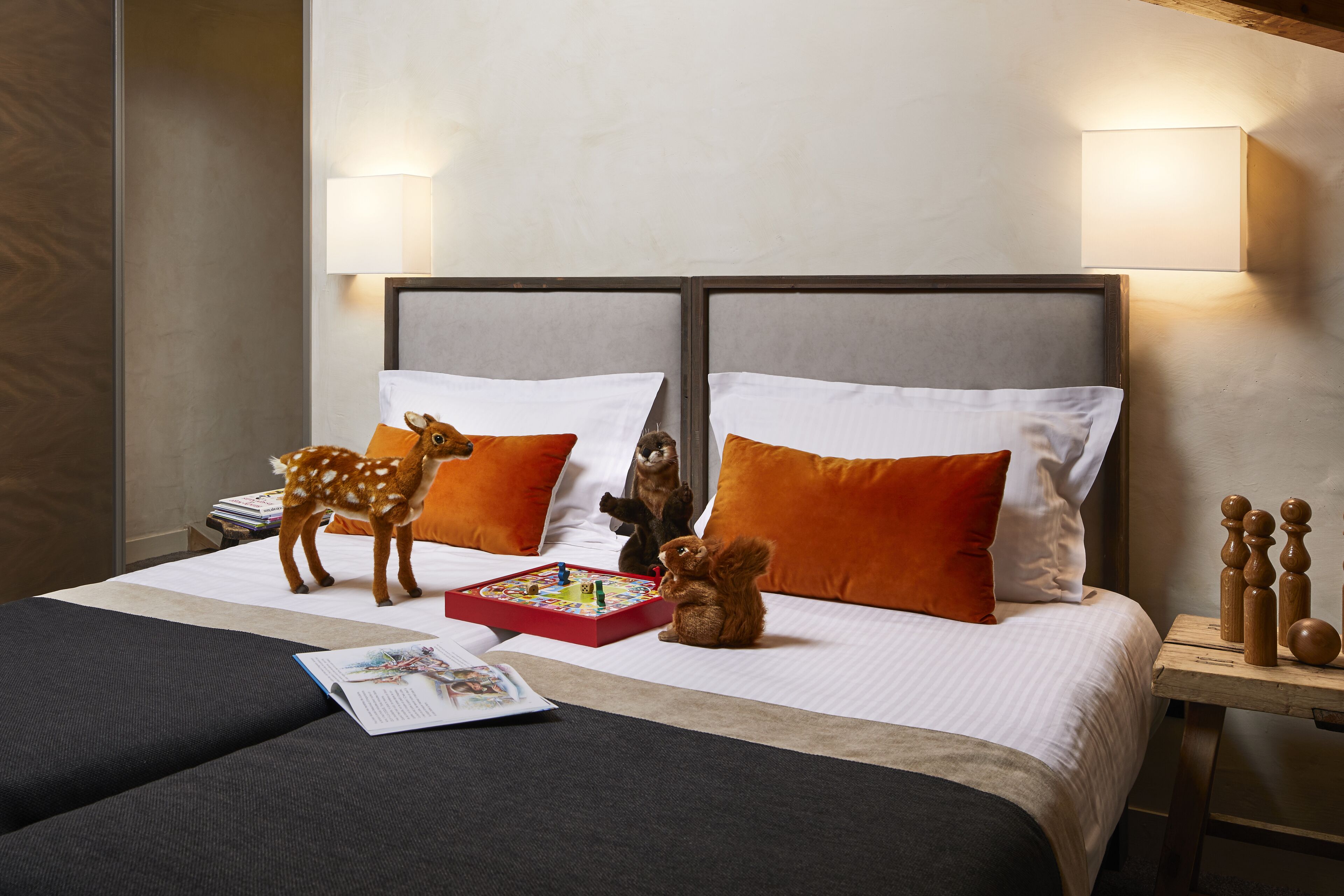 A neatly made double bed with white and orange pillows, a small table with a board game, and two stuffed animals.