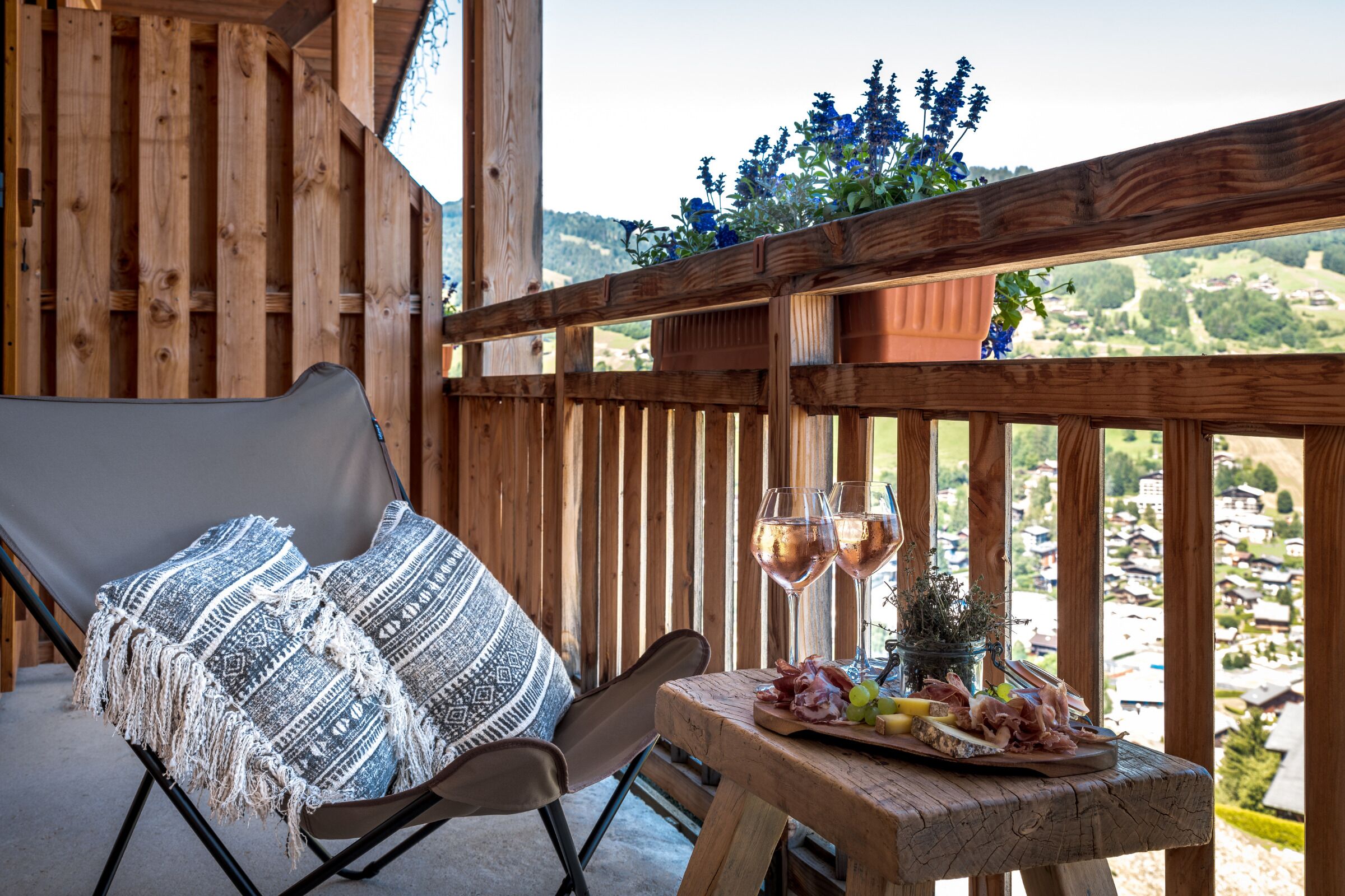 Een houten balkon met een comfortabele stoel, een klein tafeltje met een wijnglas en een bord eten, en uitzicht op een dorp.