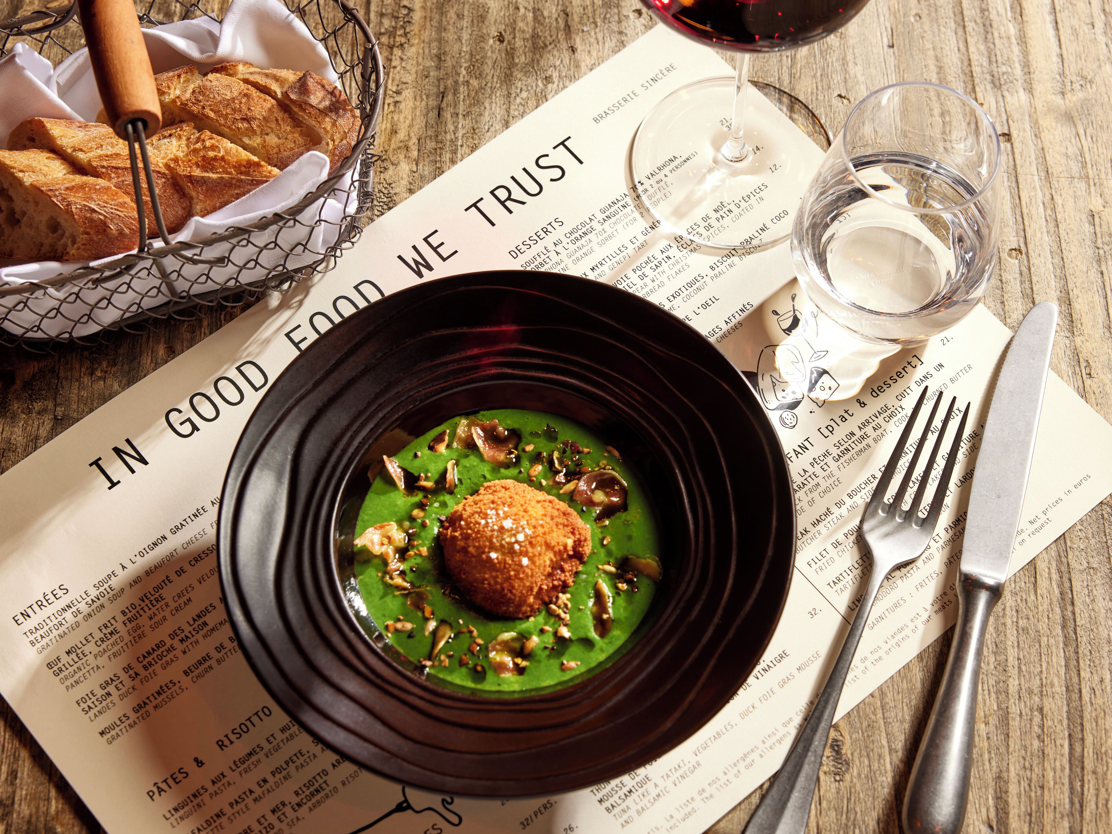 A menu with a dish of green sauce and a croquette, a basket of bread, and a glass of red wine on a wooden table.