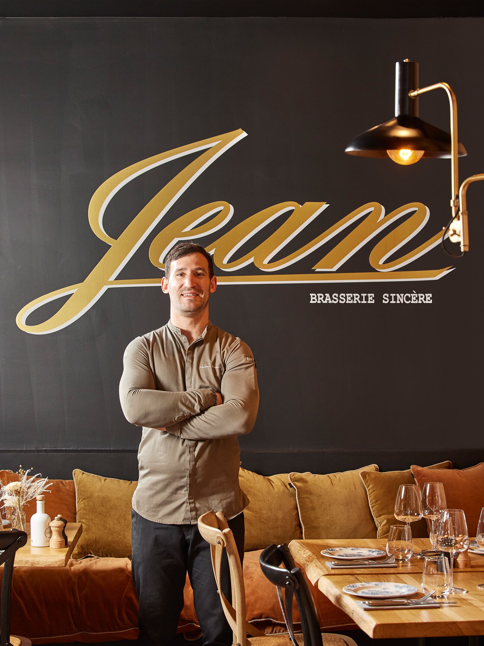 Un homme se tient dans un restaurant. Sur le mur, on peut voir une grande enseigne avec le nom « Jean ». Une table en bois, dressée avec des assiettes et des verres, se trouve devant lui. Un mur noir, orné de lettres dorées, complète le décor.