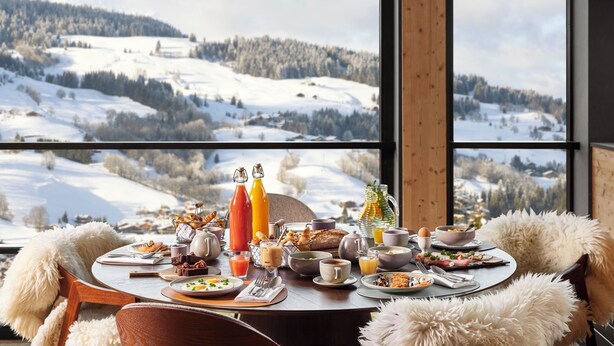 Mesa de café da manhã posta com diversos pratos e bebidas, com vista para uma paisagem nevada através de grandes janelas.
