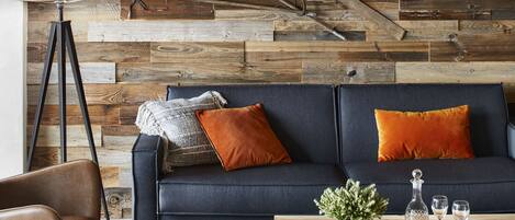A cozy living room with a dark blue sofa, orange cushions, a wooden coffee table, and a rustic wooden wall.