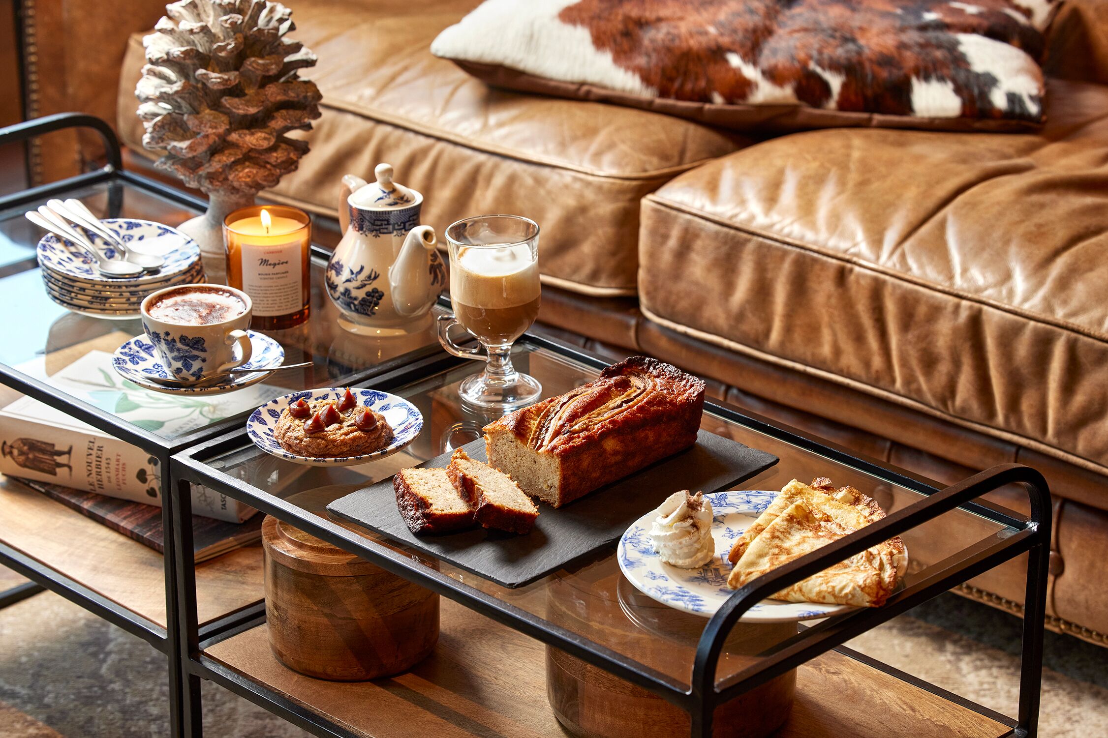 Una sala de estar acogedora con una mesa de centro de vidrio que sostiene un pan, un plato de galletas, una vela y una tetera.