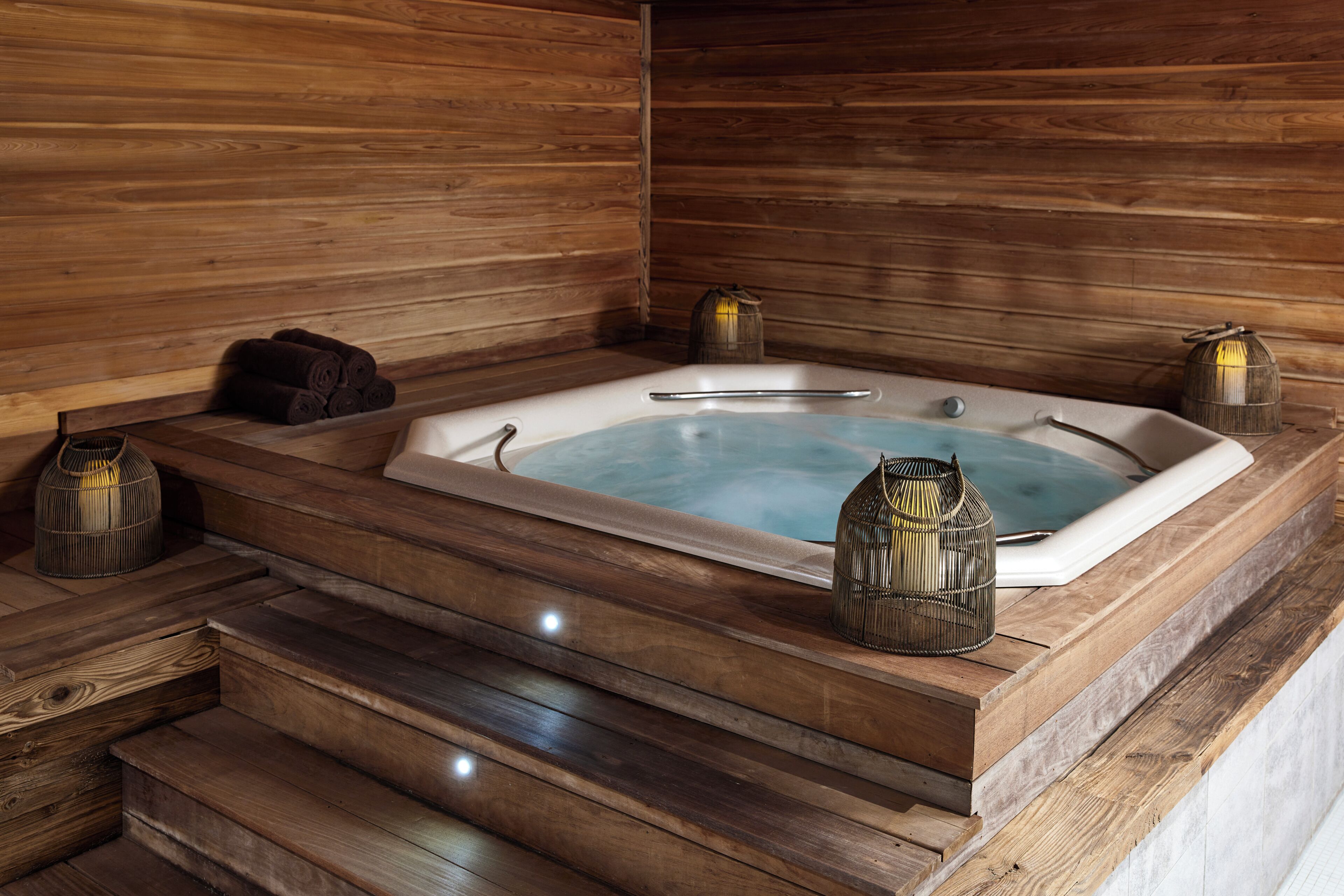 Una sauna de madera con una bañera rectangular, escalones que llevan a ella y dos faroles decorativos.