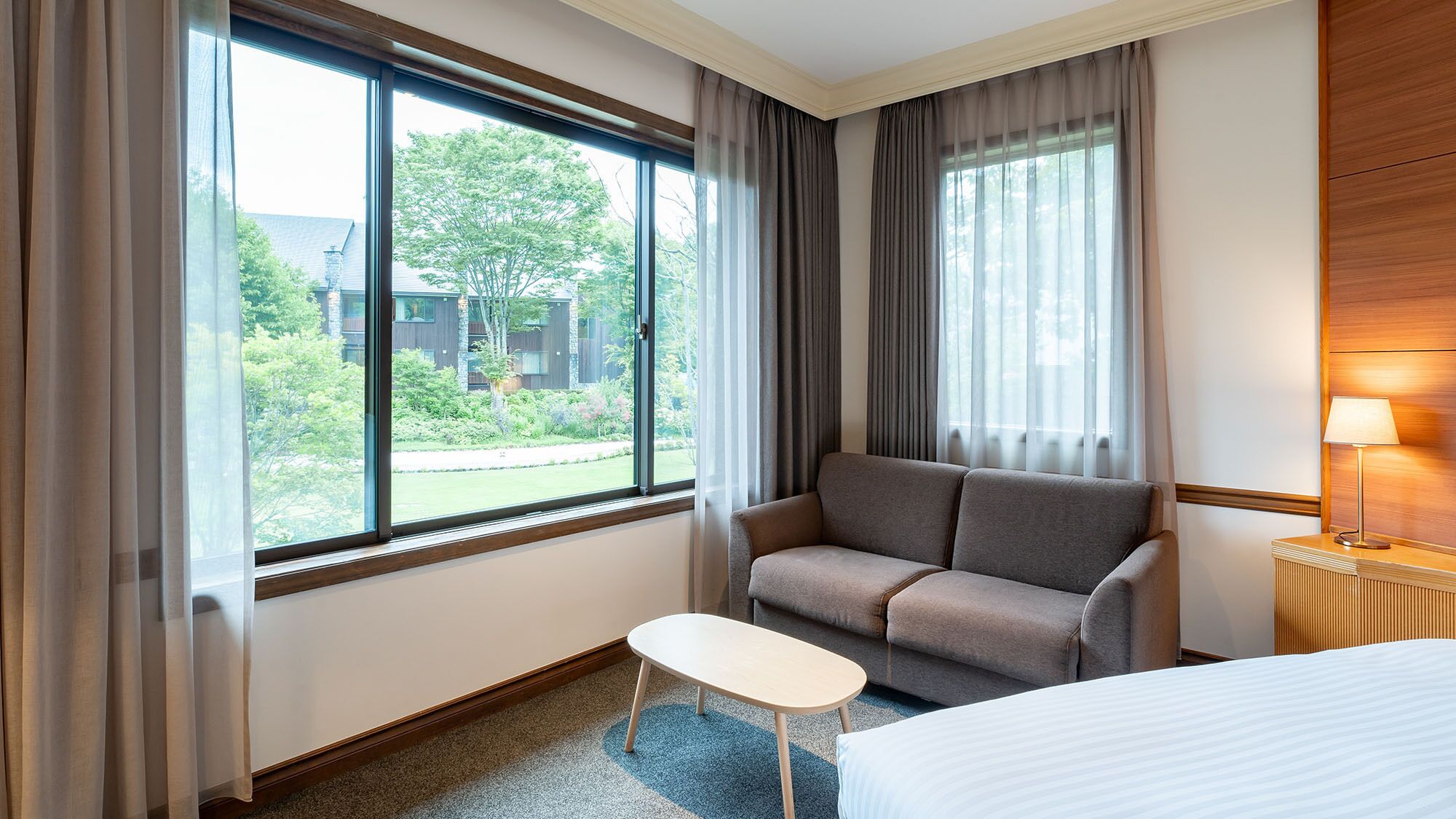 A hotel room with a sofa, a small table, a bed, and large windows with curtains.