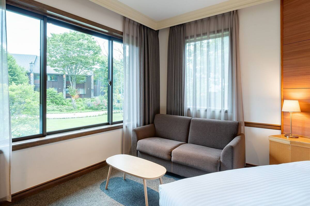 A hotel room with a sofa, a small table, a bed, and large windows with curtains.