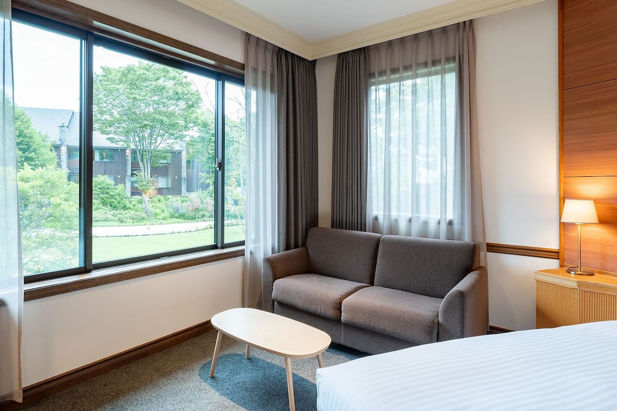 A hotel room with a sofa, a small table, a bed, and large windows with curtains.