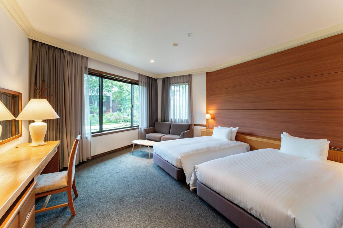 A hotel room with two beds, a desk, a chair, a sofa, and a window with curtains.
