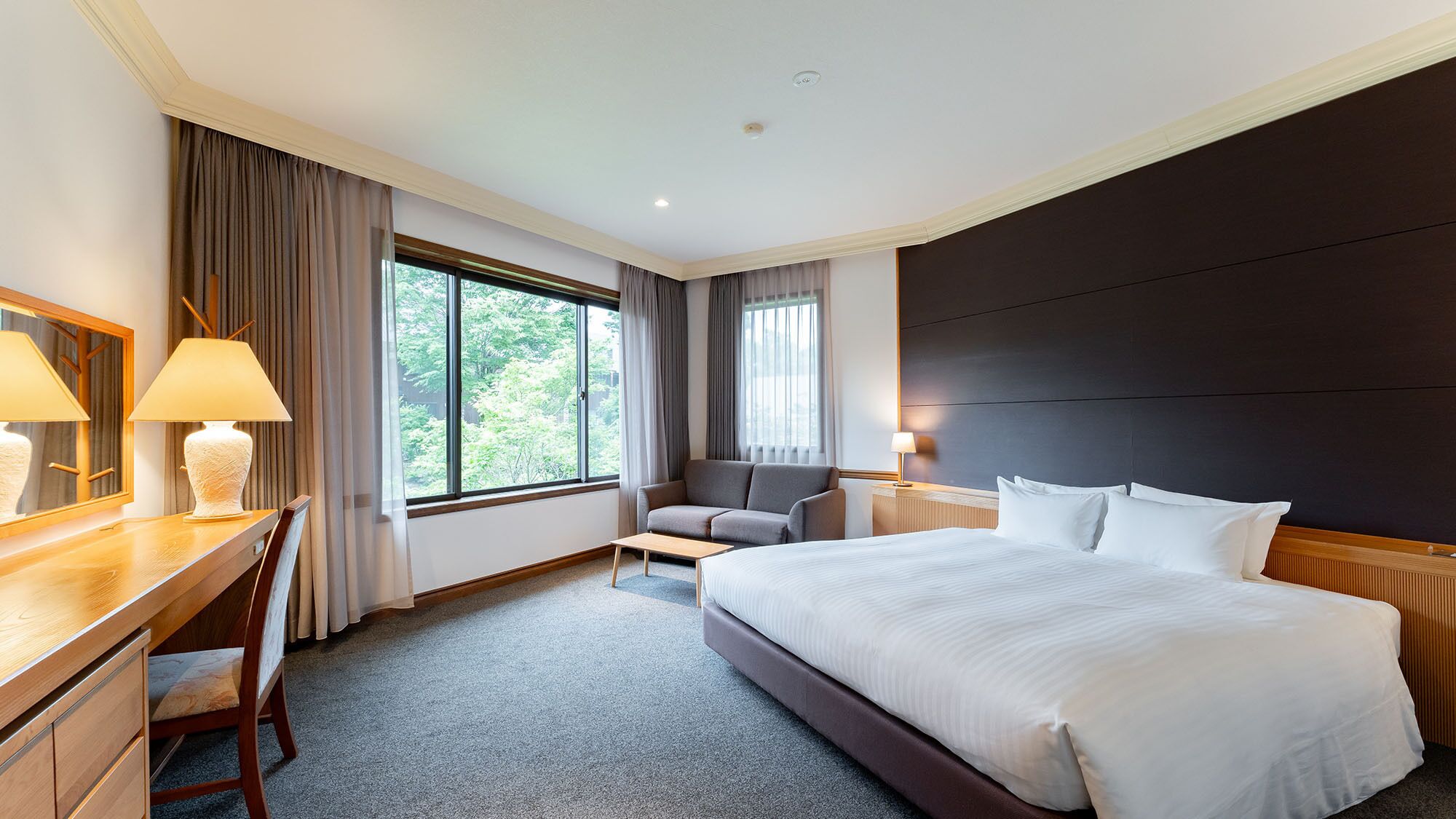 A hotel room with a large bed, a desk with a chair, a sofa, and two windows with curtains.