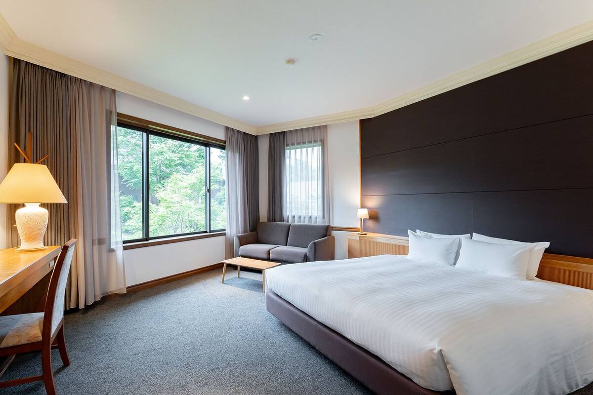 A hotel room with a large bed, a desk with a chair, a sofa, and two windows with curtains.