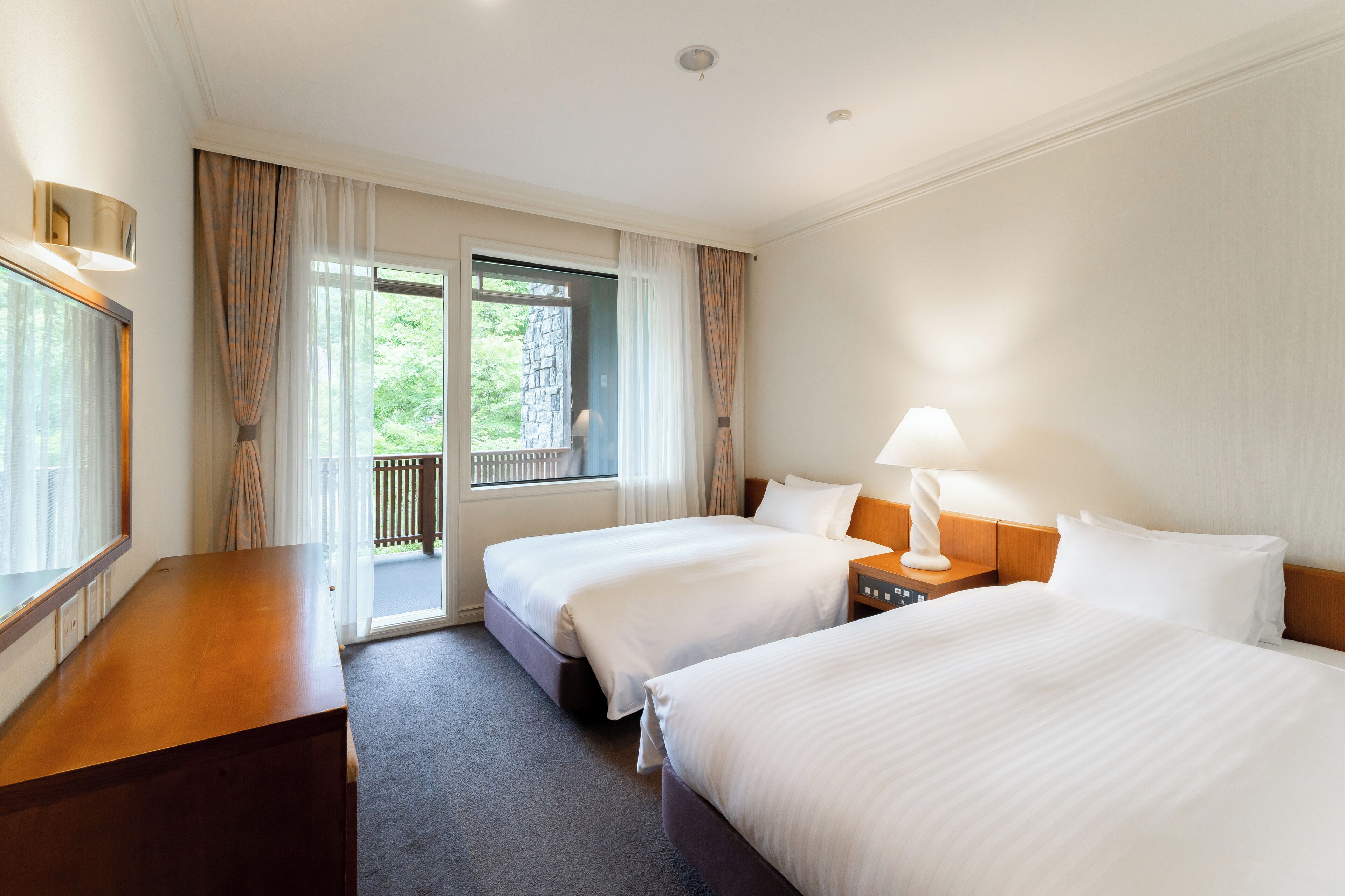 Ein Hotelzimmer mit zwei Betten, einem Holztisch, einem Nachttisch mit Lampe und Blick auf Grünflächen durch das Fenster.