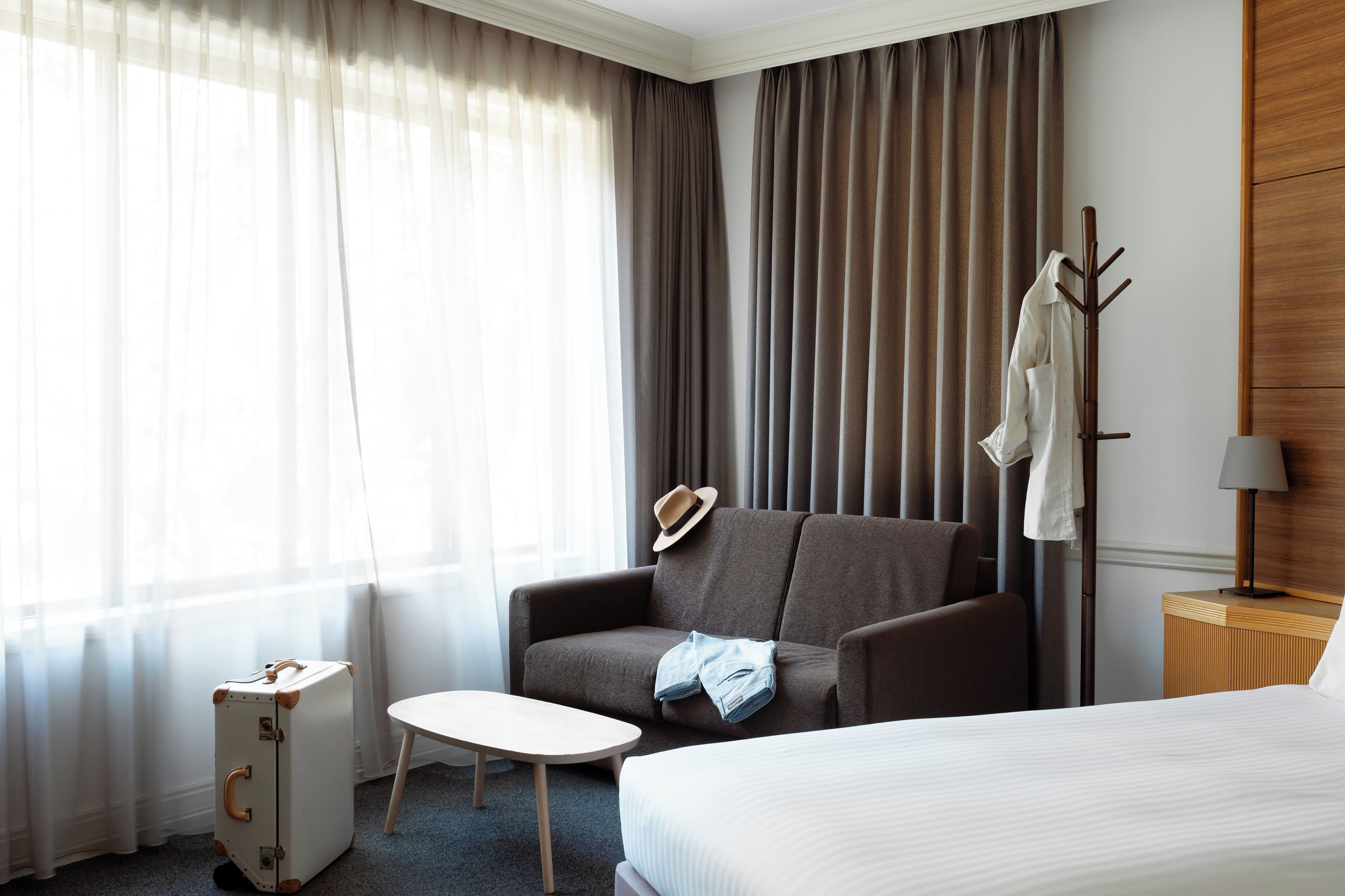 A hotel room with a bed, a sofa, a small table, a coat rack, and a suitcase.