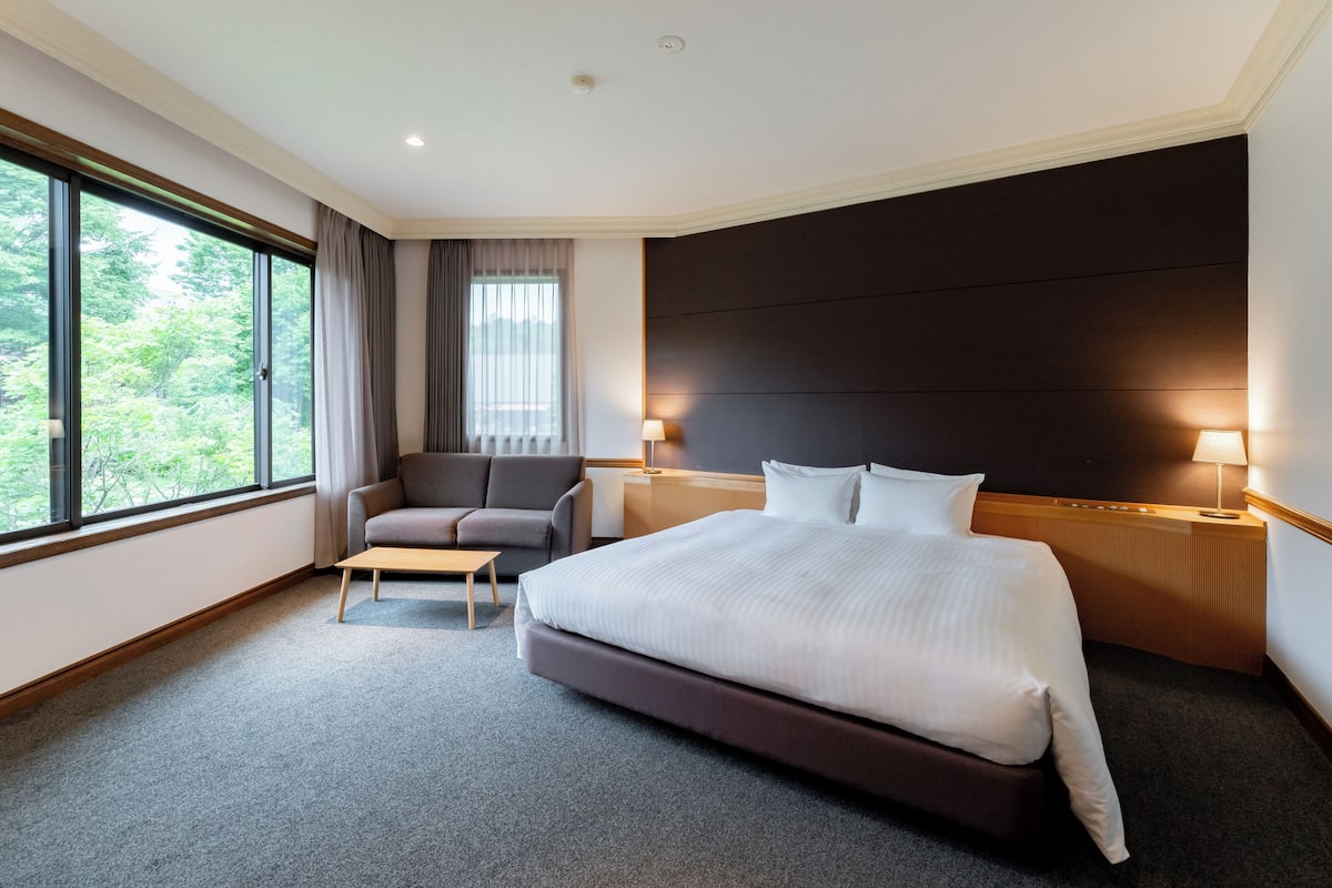 A hotel room with a large bed, a sofa, a small table, two bedside lamps, and a view of greenery through large windows.