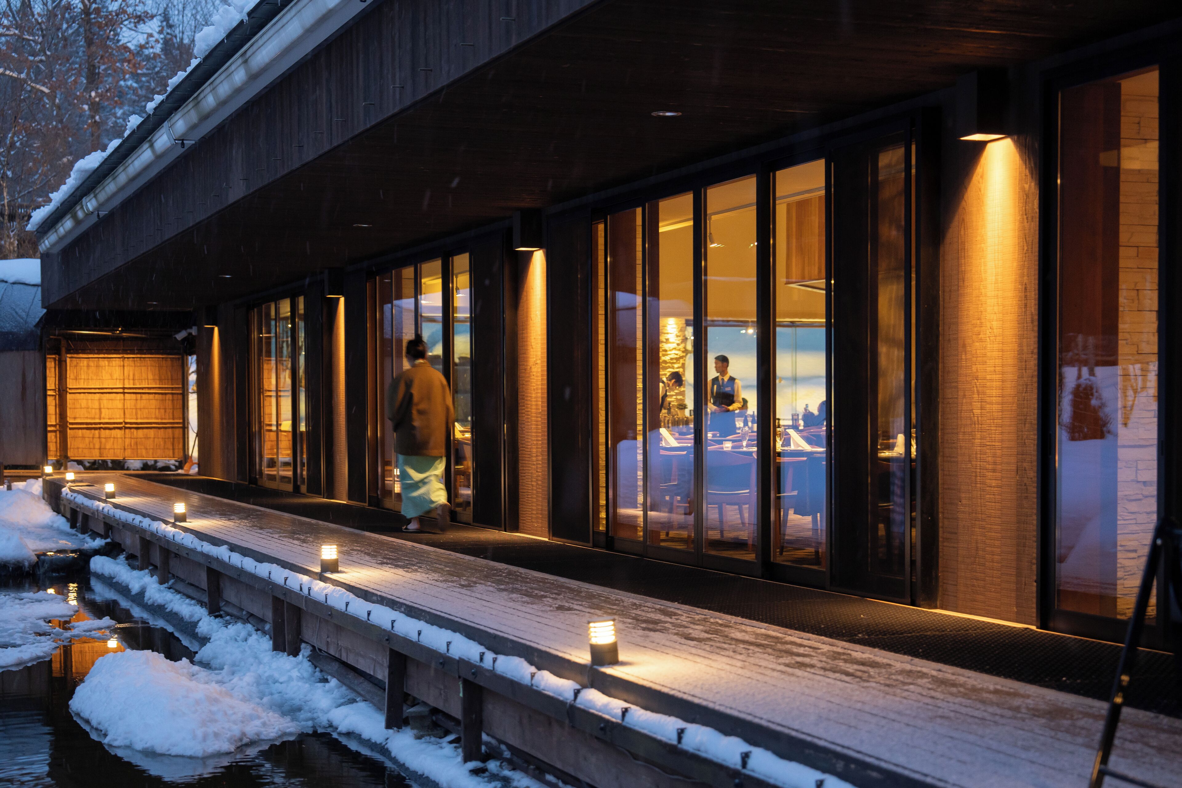 Eine Holzterrasse mit Schnee, ein Gebäude mit großen Fenstern und eine Person, die geht.