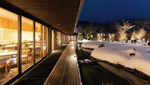 Un espacio exterior cubierto de nieve con una terraza de madera, un edificio con paredes de cristal y luces interiores, y techos nevados al fondo.