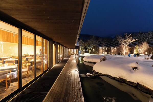 Un espacio exterior cubierto de nieve con una terraza de madera, un edificio con paredes de cristal y luces interiores, y techos nevados al fondo.