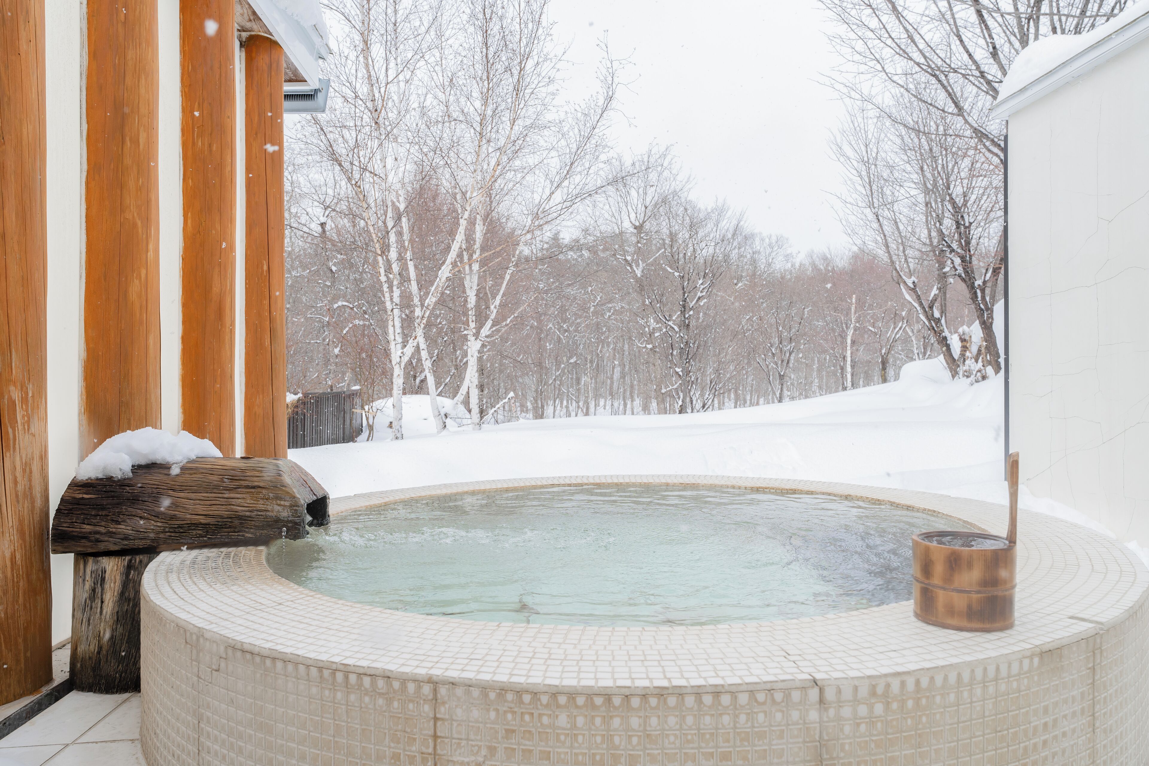 Un jacuzzi con un banco de madera, rodeado de árboles cubiertos de nieve y un edificio blanco.
