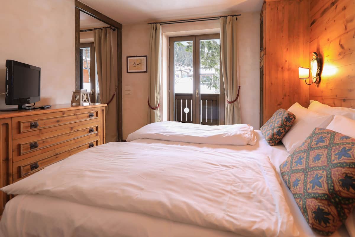 Quarto com cômoda de madeira, televisão, cama com roupa de cama branca, almofadas estampadas e janela com cortinas.