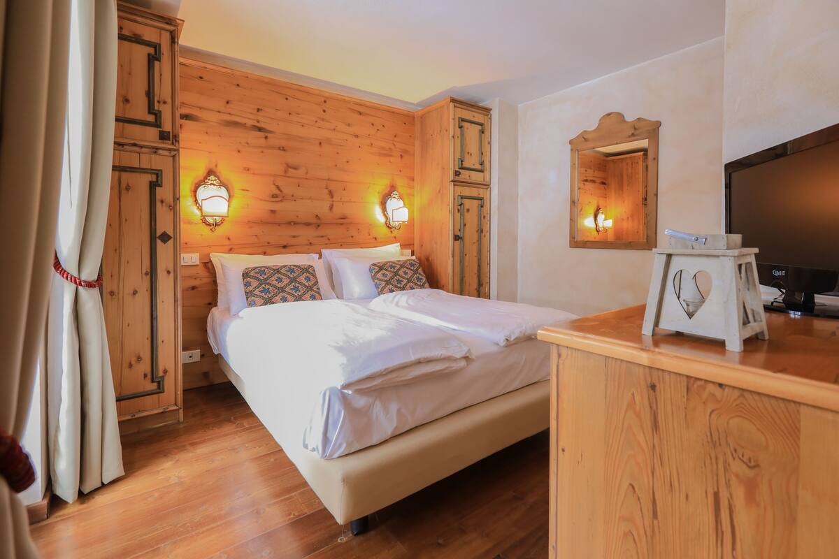 Quarto com cabeceira de madeira, duas luminárias de cabeceira, cama com roupa de cama branca, penteadeira de madeira e uma televisão sobre um suporte.