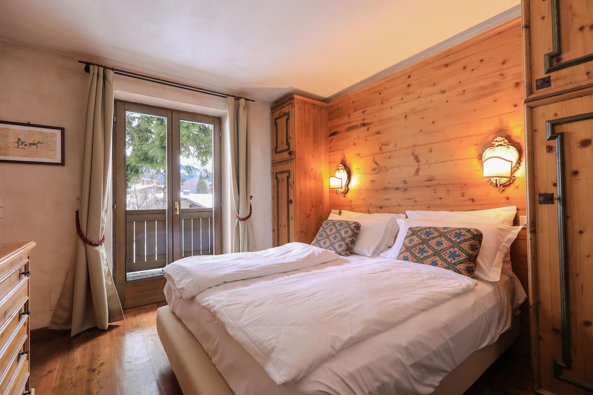 Quarto com cabeceira de madeira, duas camas com roupa de cama branca, cómoda de madeira e janela com cortinas.