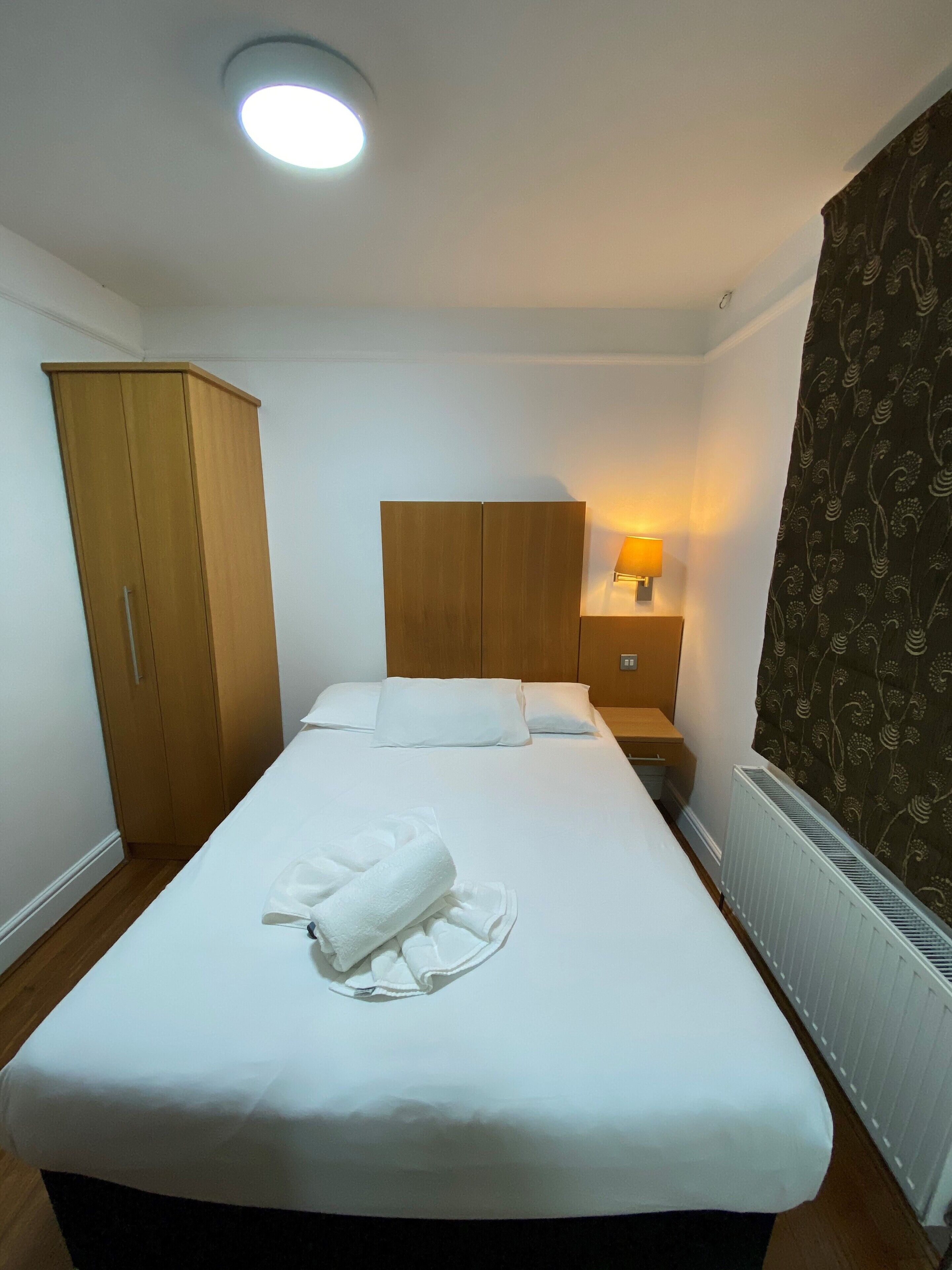 A neatly made bed with a white duvet and a rolled towel, a wooden wardrobe, a bedside table with a lamp, and a radiator.