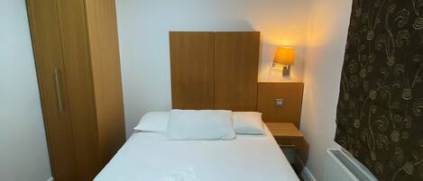 Ein ordentlich bezogenes Bett mit weißer Daunendecke und zusammengerolltem Handtuch, ein Holzschrank, ein Nachttisch mit Lampe und ein Heizkörper.