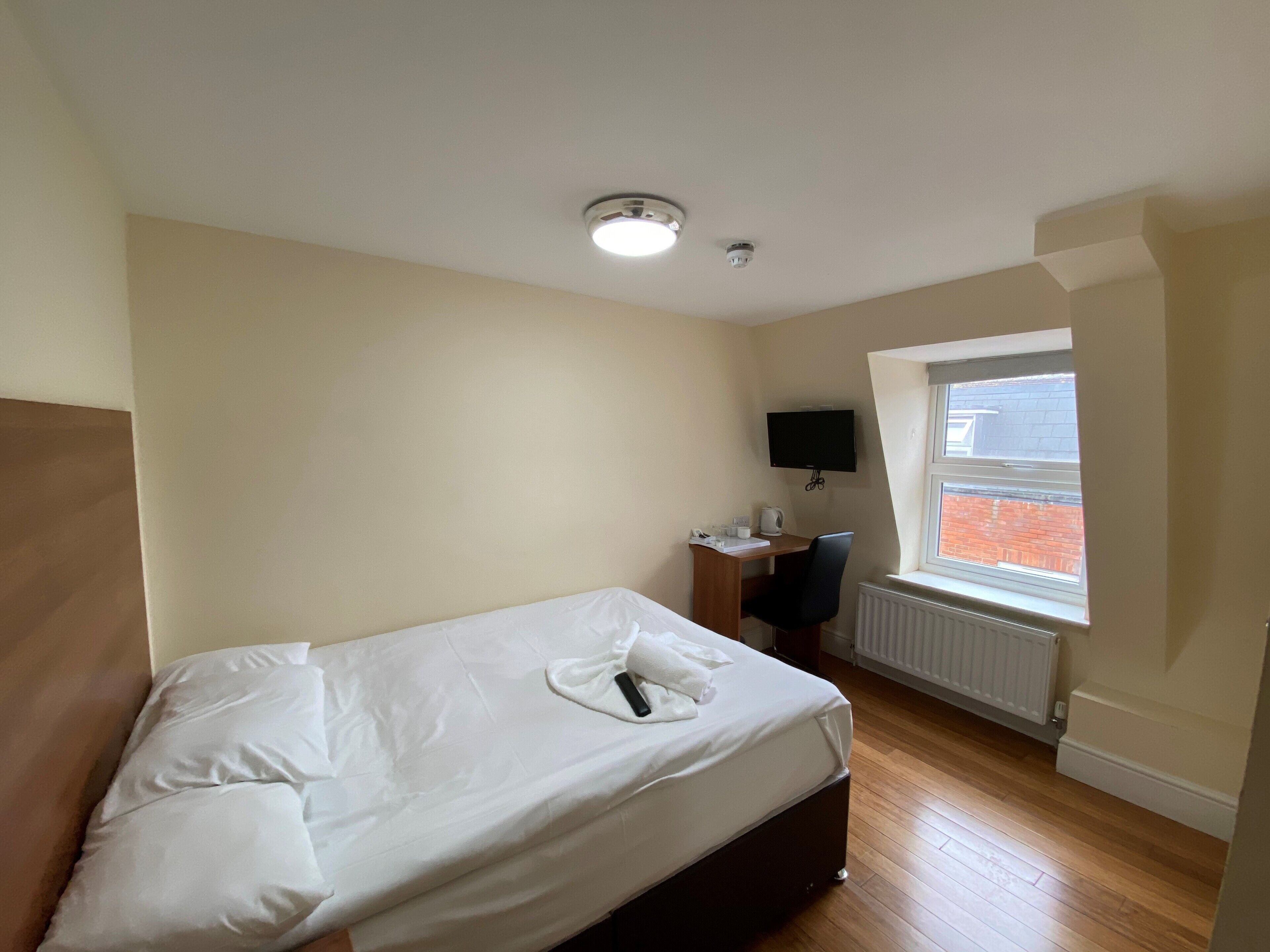 Un dormitorio con cama, escritorio, silla, televisor y ventana con vista a un edificio de ladrillo.