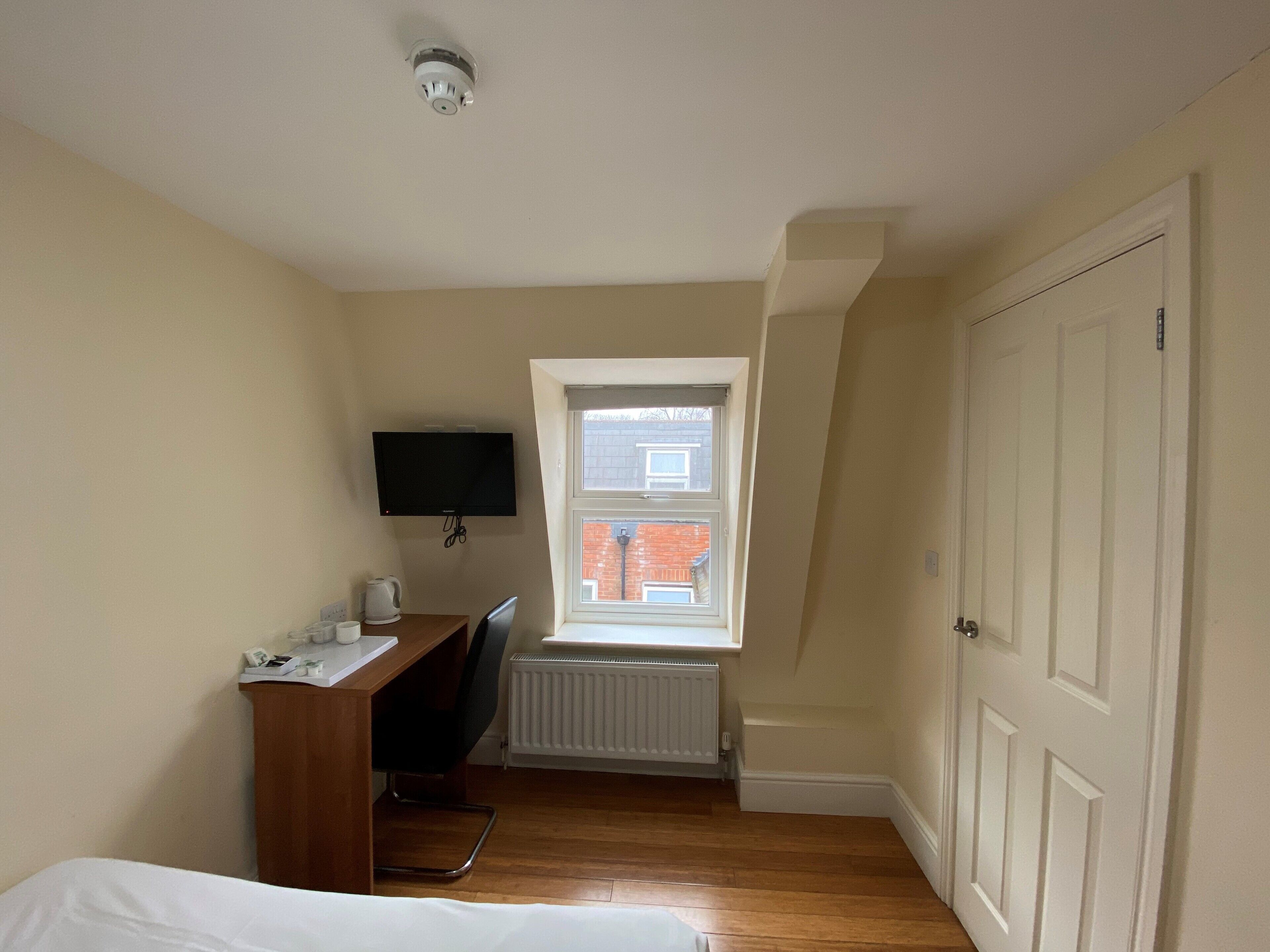 Quarto pequeno e organizado com escrivaninha de madeira, cadeira, cama, radiador, janela com vista para um prédio de tijolos e porta fechada.