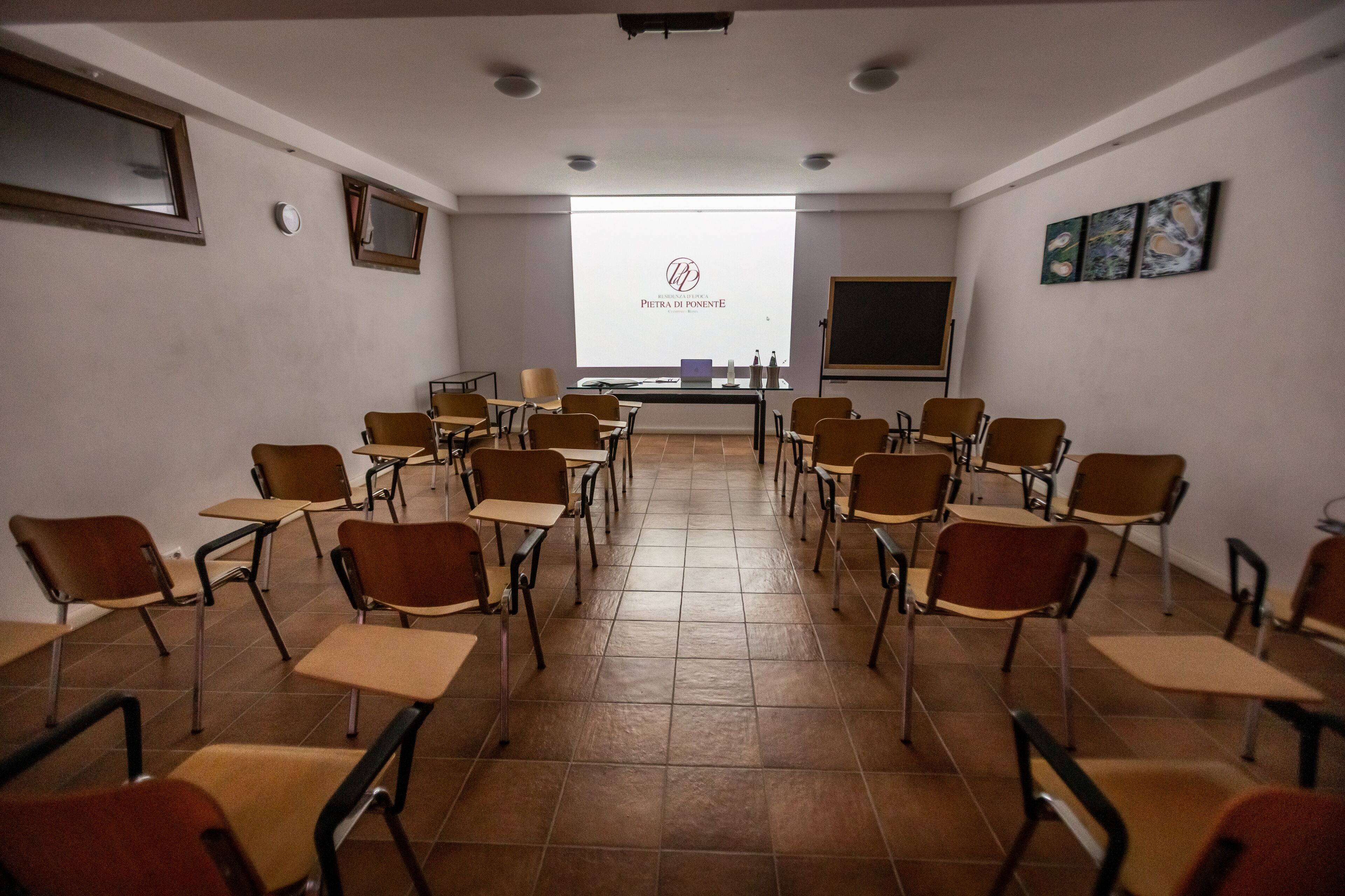 Uma sala de aula com fileiras de mesas e cadeiras, uma tela de projetor e um quadro negro.