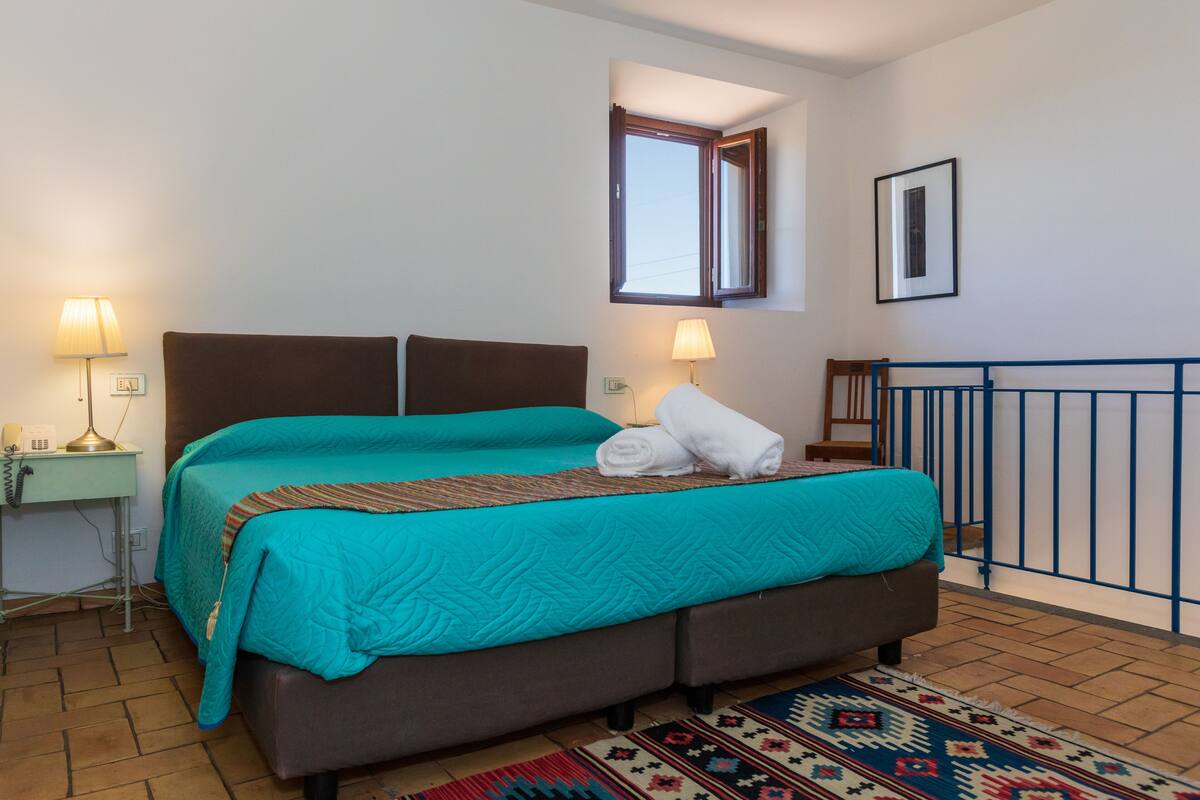 Ein ordentlich eingerichtetes Schlafzimmer mit einem Bett, zwei Nachttischlampen, einem kleinen Schreibtisch mit Lampe, einem Fenster und einem Bild an der Wand.