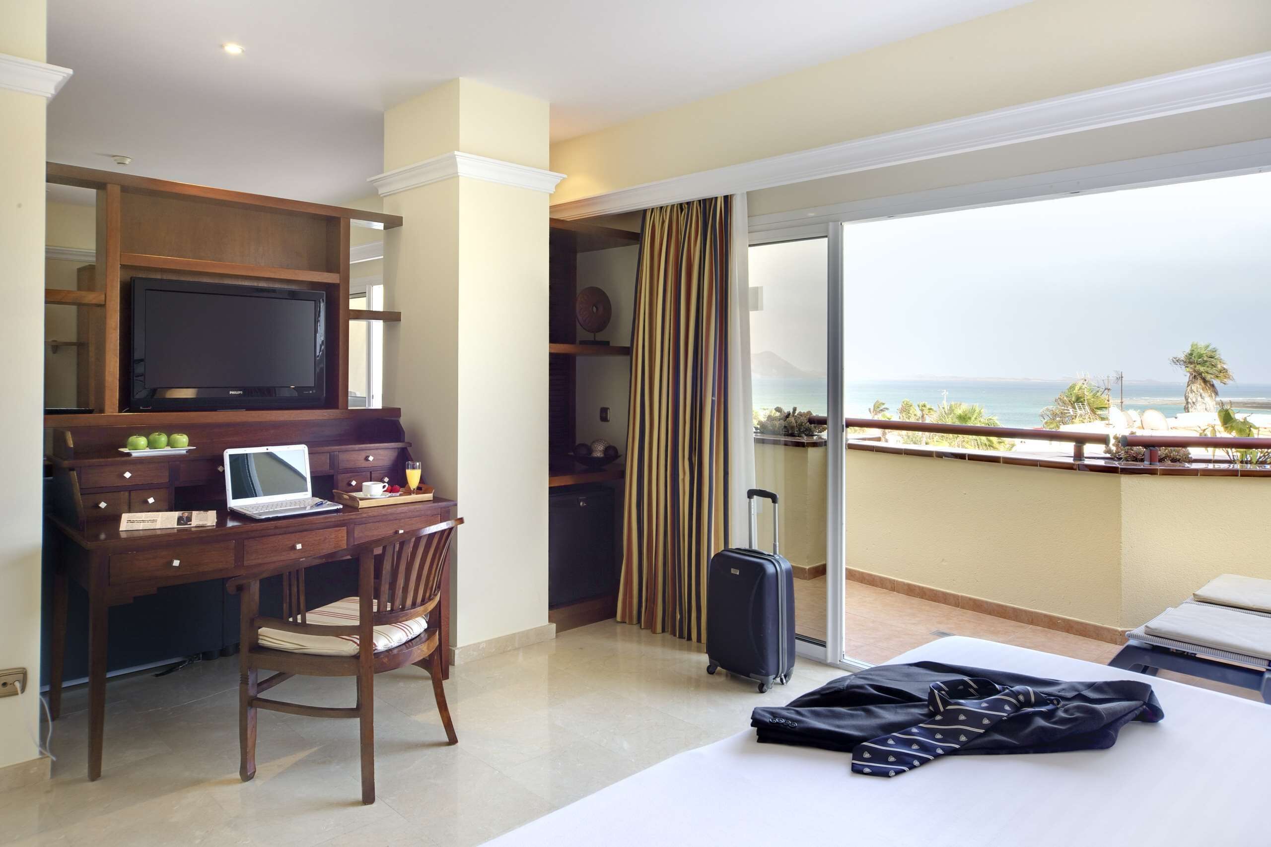 Habitación de hotel con escritorio, computadora portátil, silla, cama, maleta y un balcón con vista al mar.