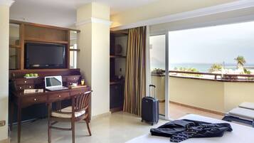 Habitación de hotel con escritorio, computadora portátil, silla, cama, maleta y un balcón con vista al mar.
