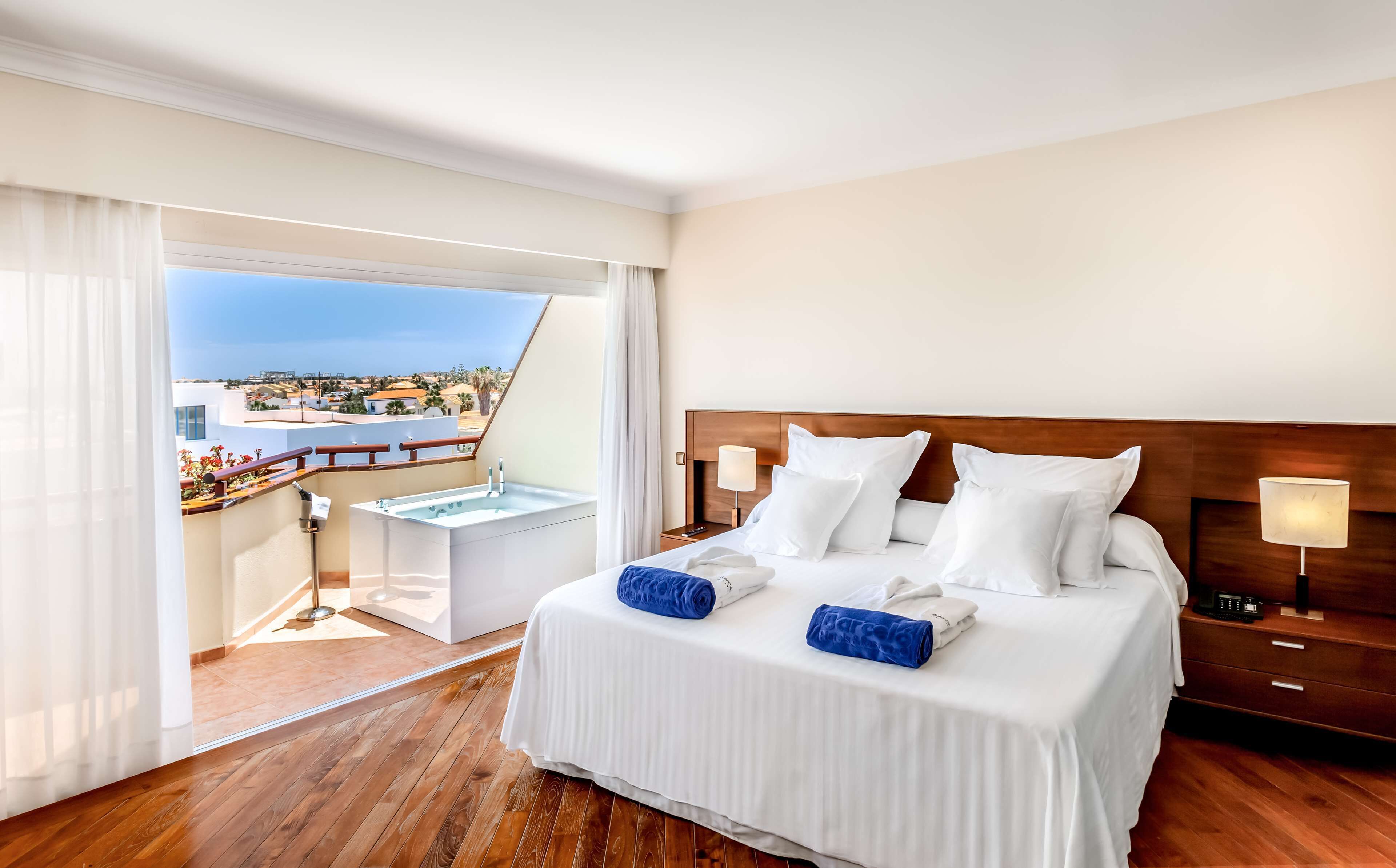 A hotel room with a large bed, a wooden floor, a balcony with a view of the city, and a small table with a lamp.