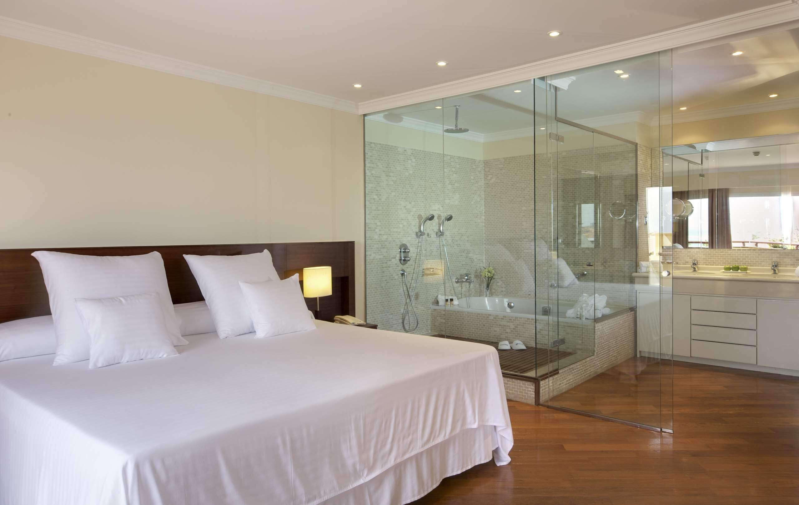 Une chambre d’hôtel moderne dotée d’un grand lit, d’une douche séparée par des parois en verre et d’une salle de bain à l’aménagement ouvert.