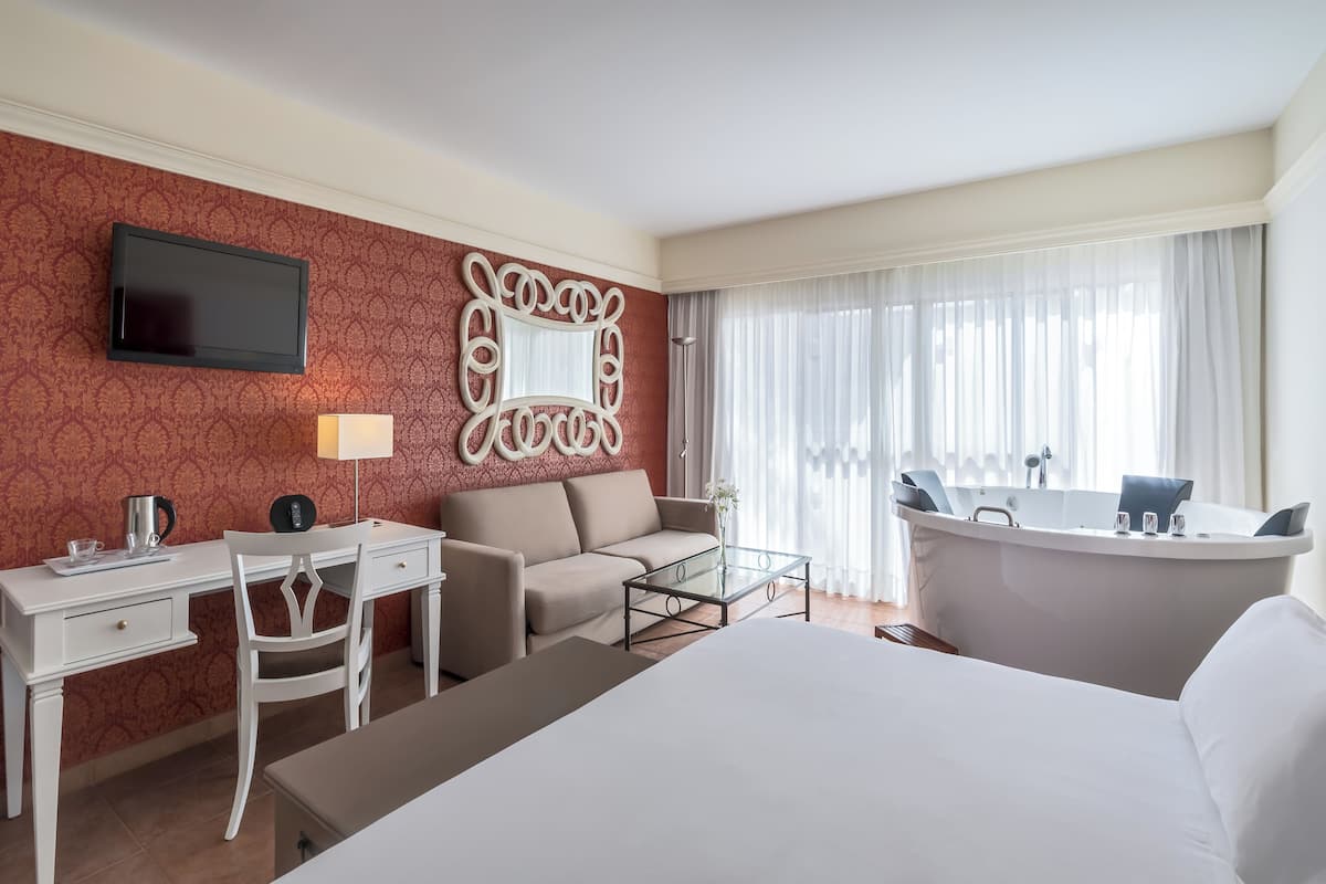 A hotel room with a bed, a desk with a chair, a sofa, a glass coffee table, a large mirror, a flat-screen TV, a bathtub, and a window with sheer curtains.