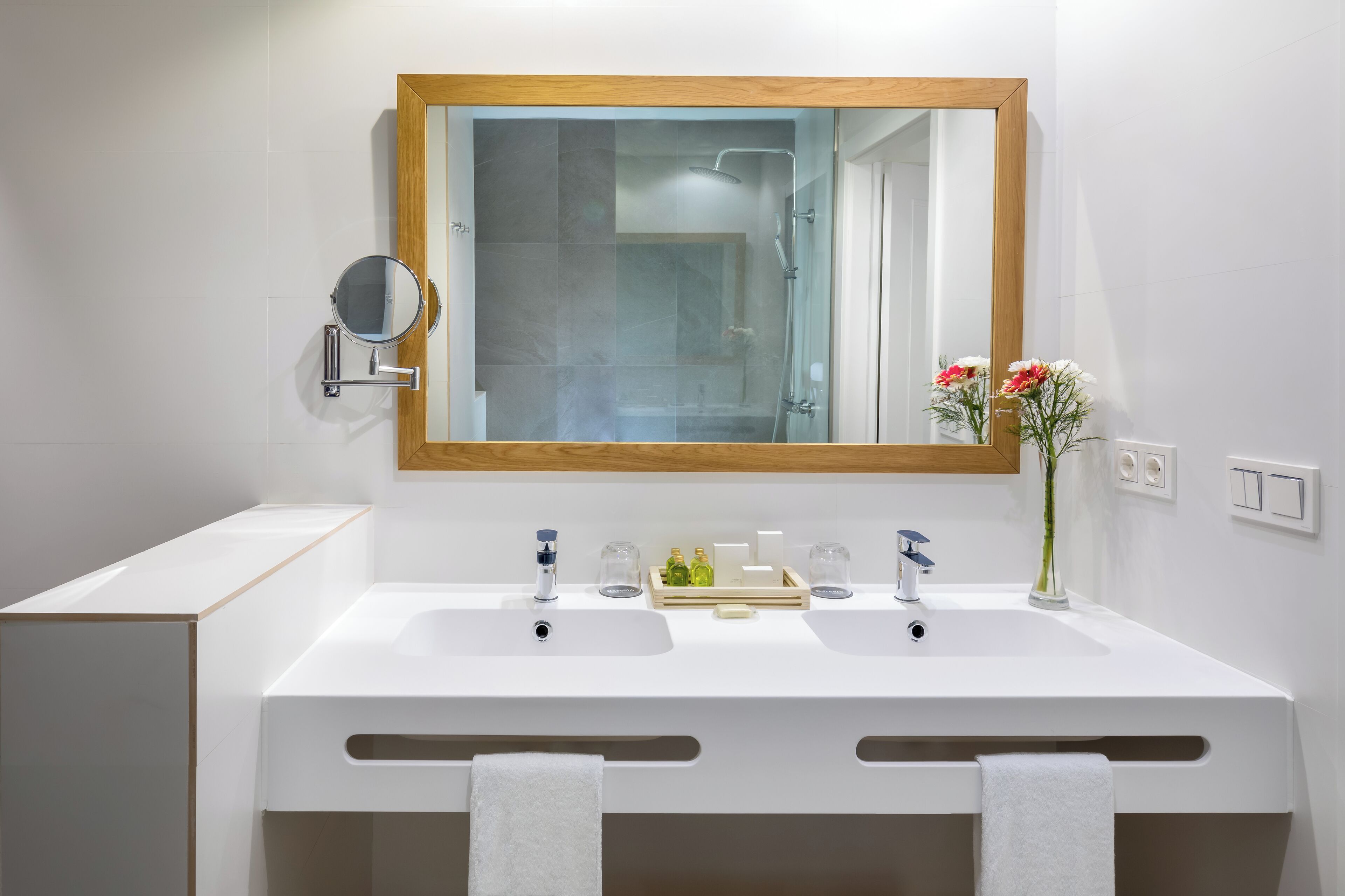 Une salle de bain moderne avec un grand miroir, deux lavabos et une douche. On y trouve une petite étagère avec des articles de toilette et un vase rempli de fleurs.