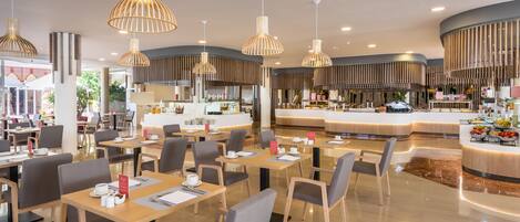 A modern restaurant with wooden tables and chairs, hanging pendant lights, and a central counter with a variety of food items.