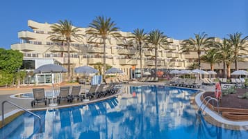 Une piscine entourée de palmiers, agrémentée de chaises longues et de parasols, et bordée d’un immeuble à plusieurs étages doté de balcons.