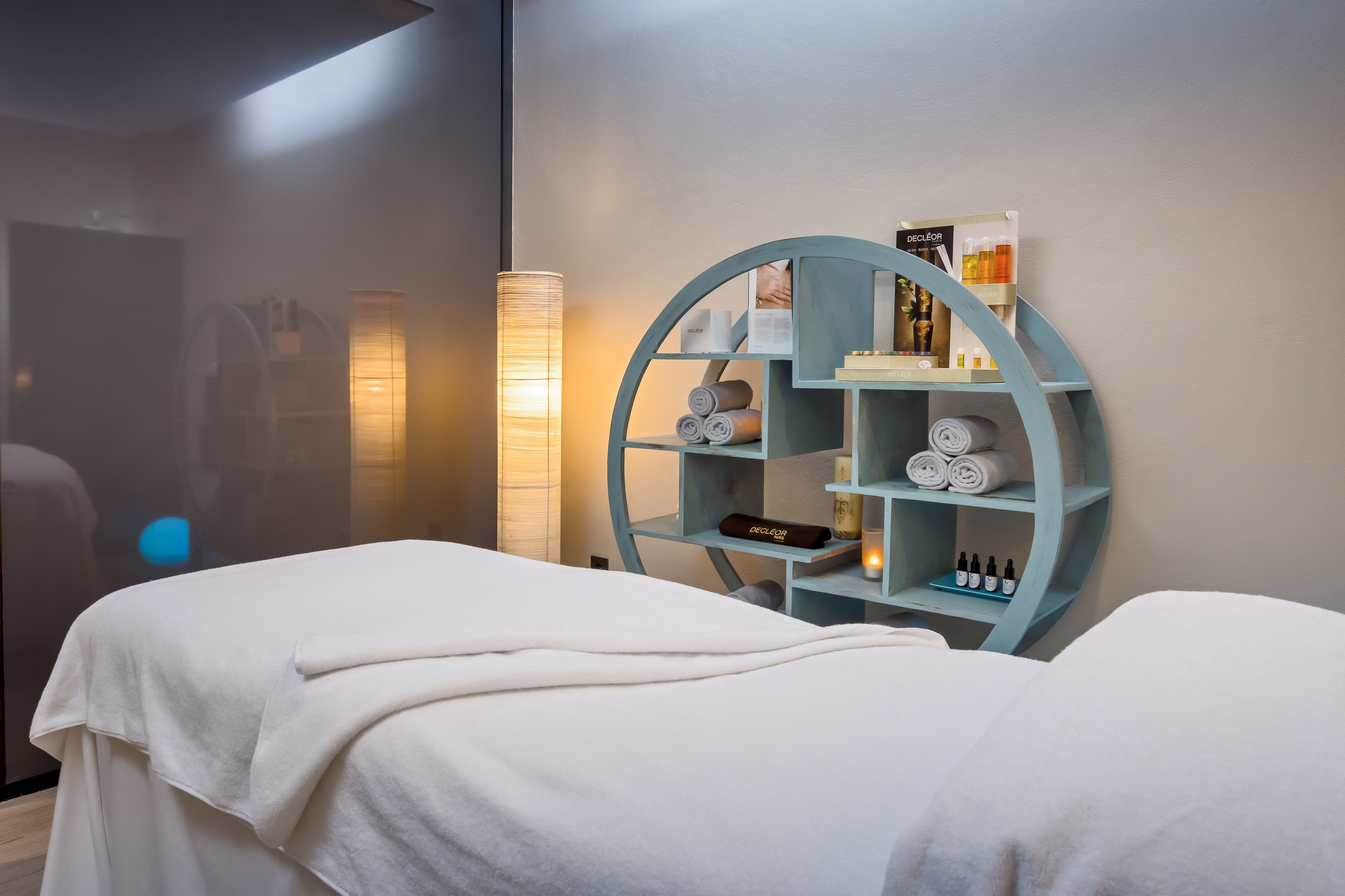 Un cuarto de spa con una camilla de masaje, un estante azul con toallas y velas, y una lámpara fijada a la pared.
