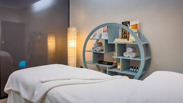 Un cuarto de spa con una camilla de masaje, un estante azul con toallas y velas, y una lámpara fijada a la pared.
