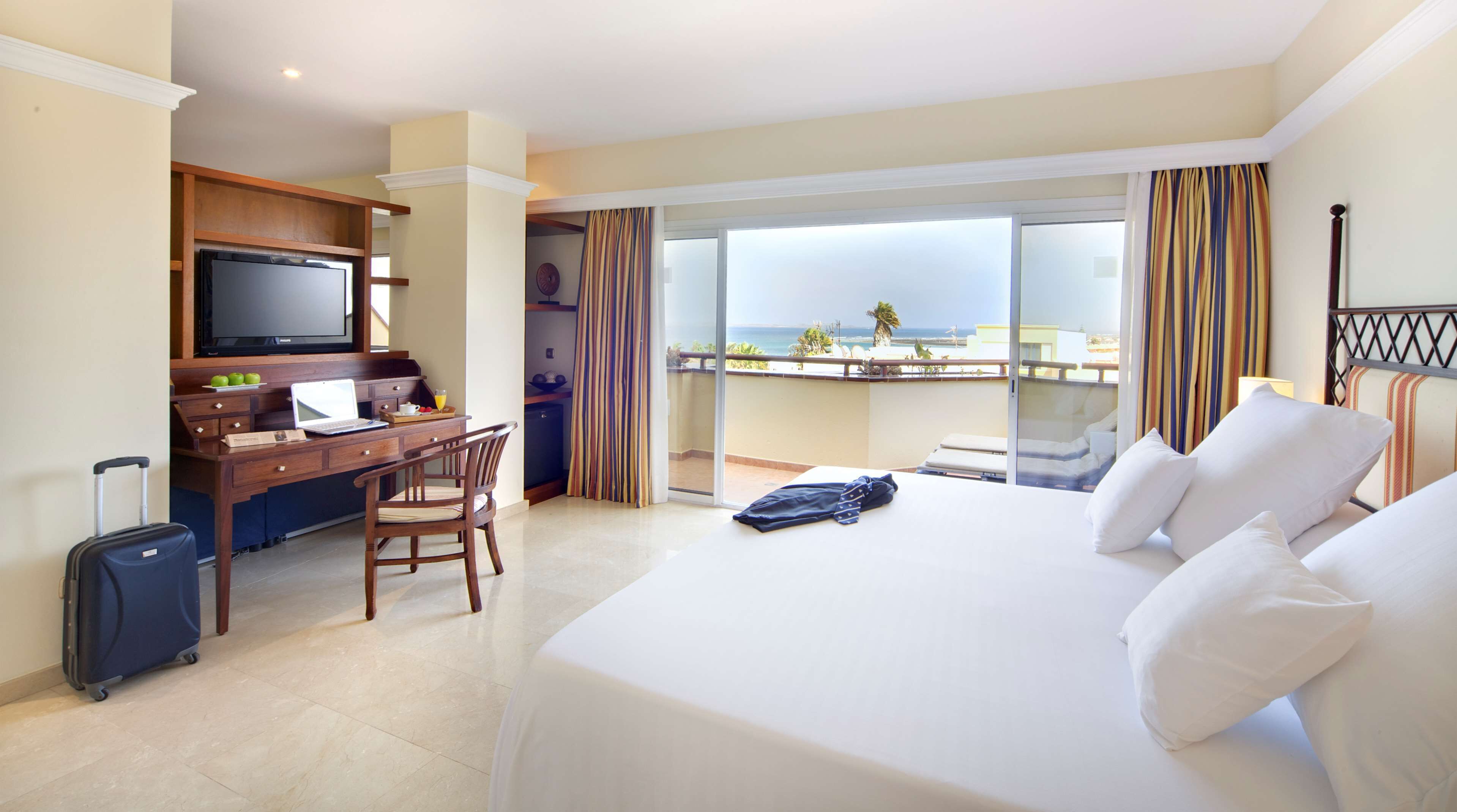 A hotel room with a bed, a desk with a laptop, a TV, and a view of the ocean.