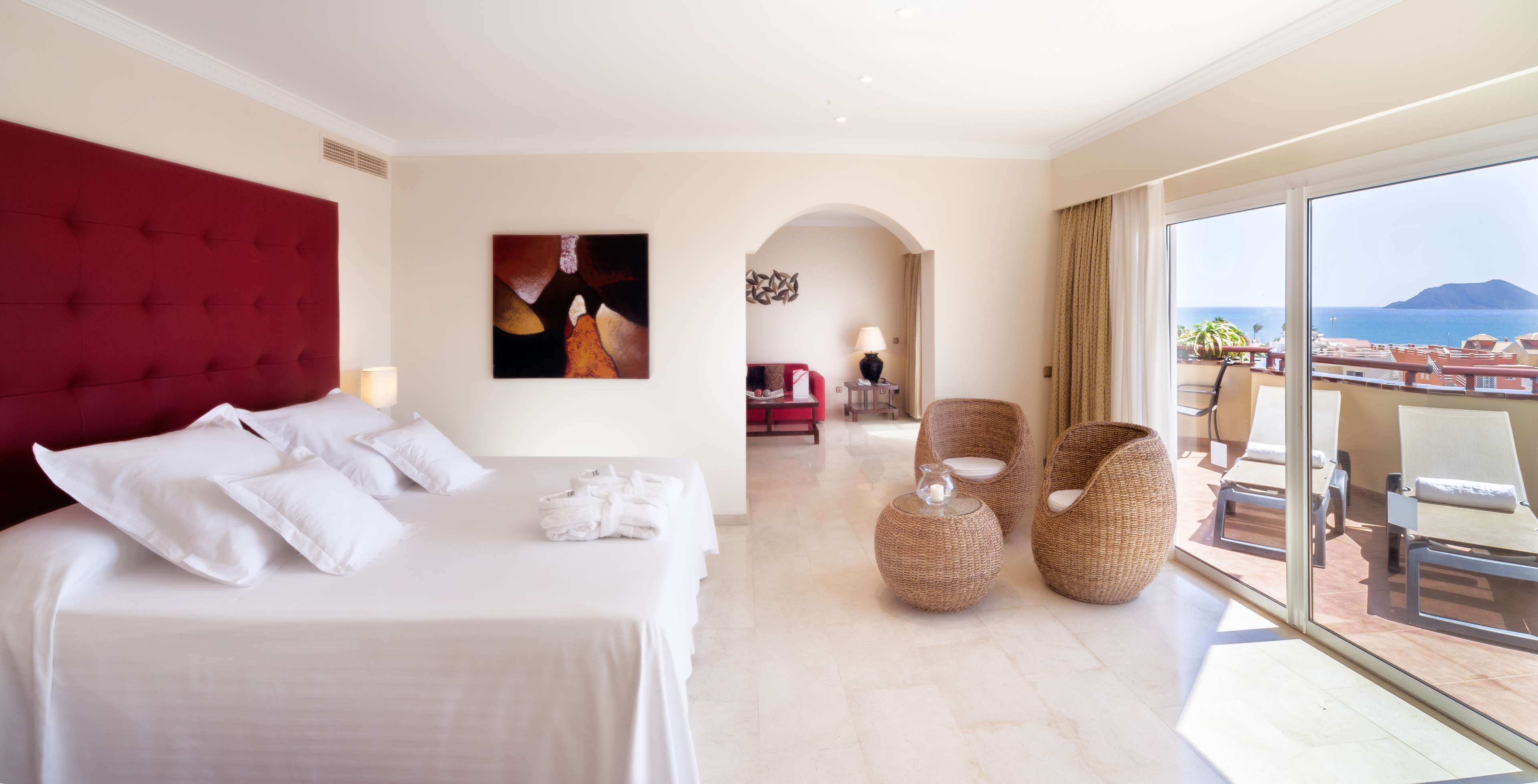 A hotel room with a large bed, a view of the sea, wicker chairs, and a balcony with sun loungers.