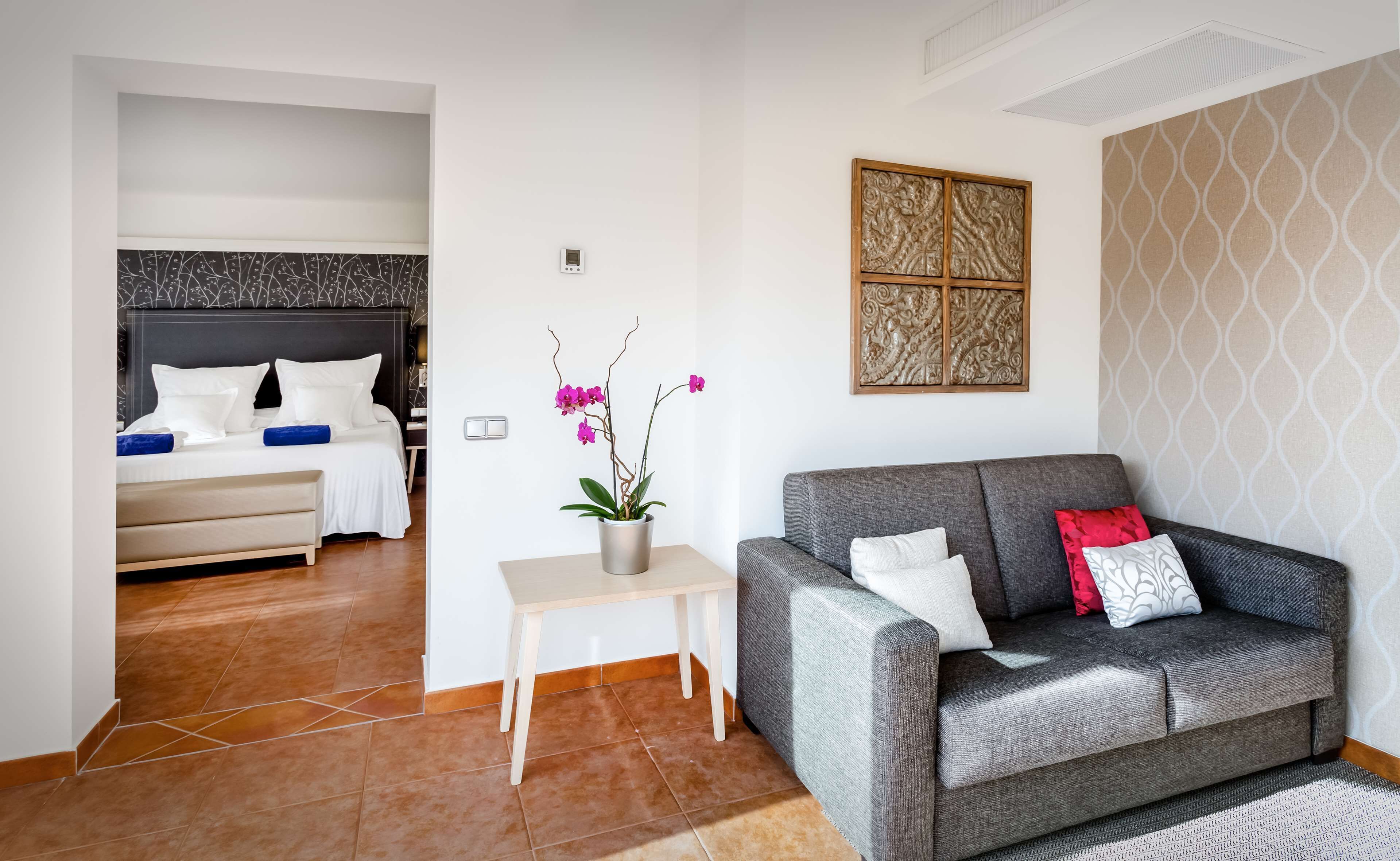 A hotel room with a bed, a grey sofa, a small table with a potted orchid, and a decorative panel on the wall.