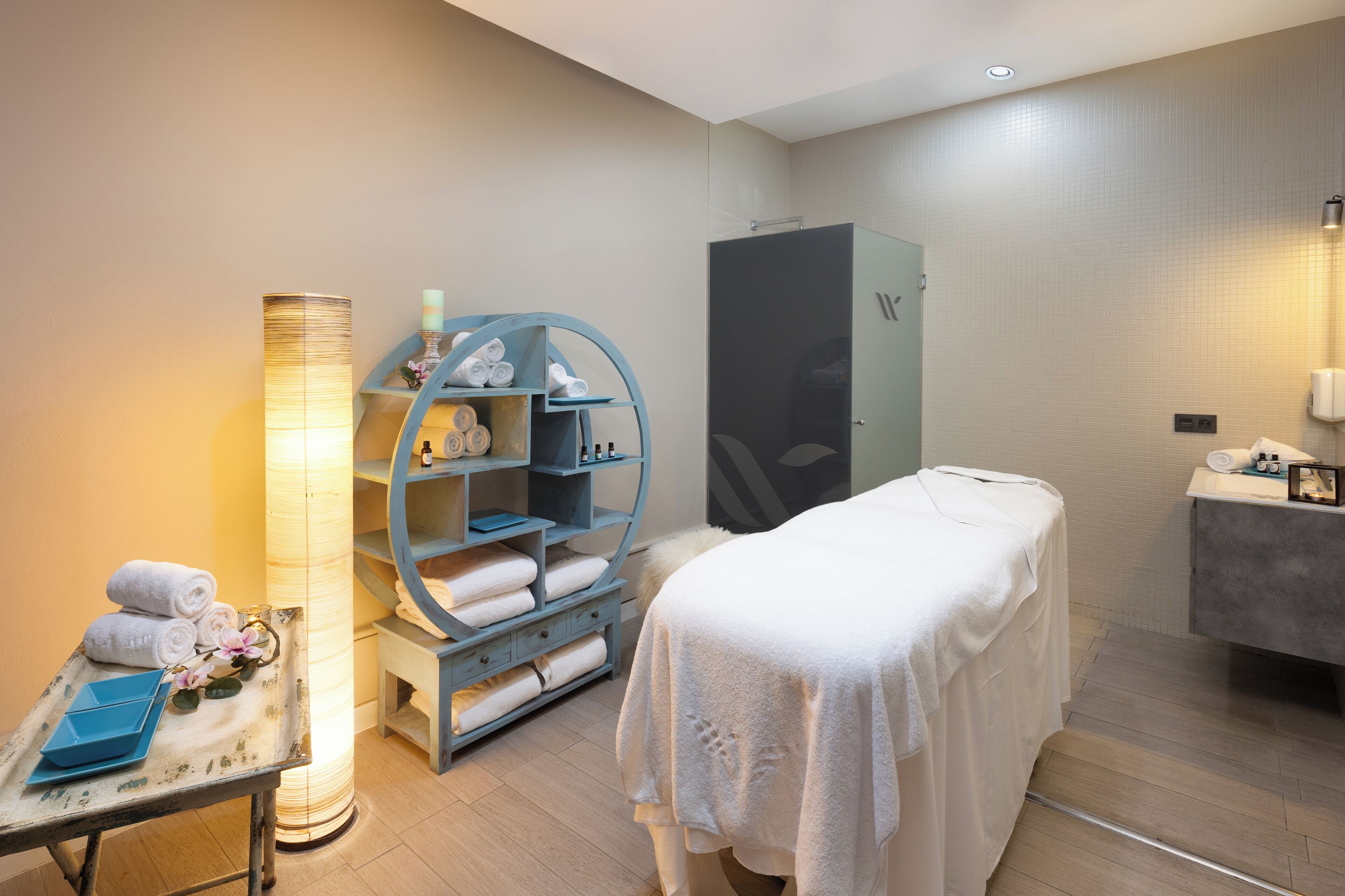 A modern spa room with a massage table, a blue shelving unit with towels, a large floor lamp, and a wooden cabinet.