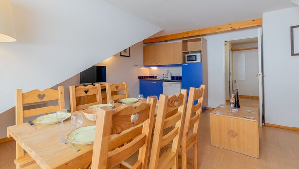 A dining area with a wooden table set for four, surrounded by chairs. A microwave and a small refrigerator are visible in the background. - Madame Vacances Residence Le Parc des Airelles (Les Orres)