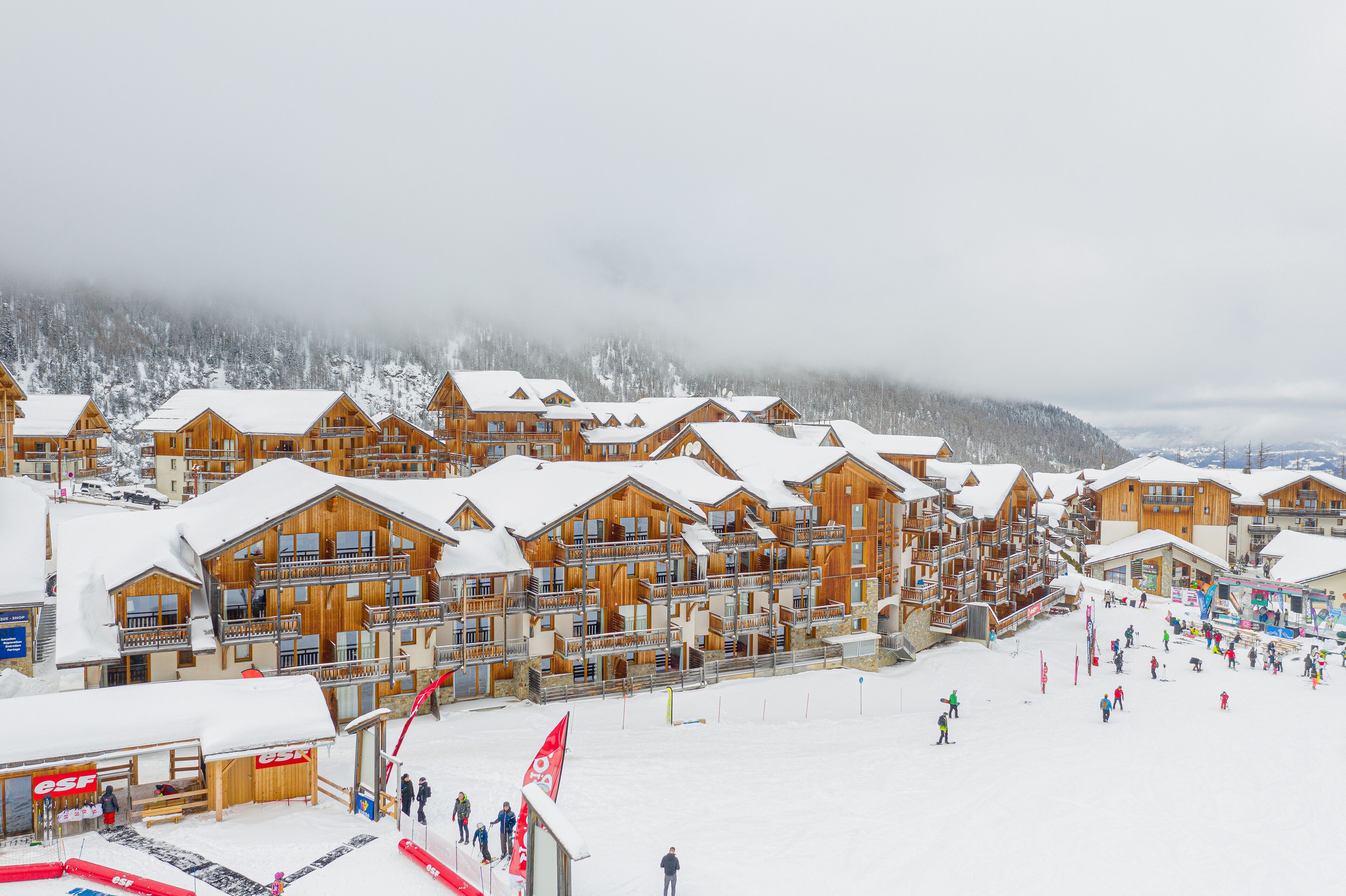 A row of wooden chalet-style buildings with balconies, a ski slope in the foreground, and a forested mountain in the background.