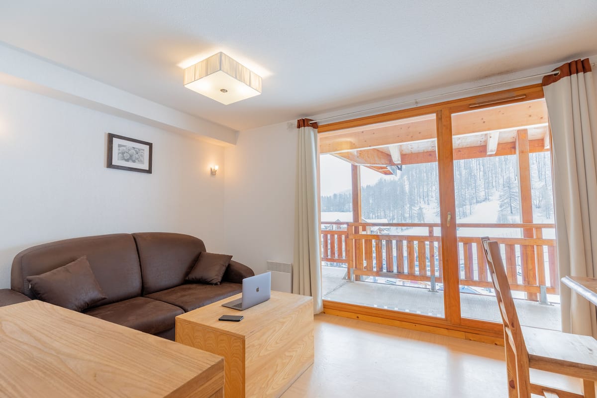 A modern living room with a brown leather sofa, a wooden coffee table, and a laptop. There is a large window with a view of a snowy landscape and a wooden deck outside.