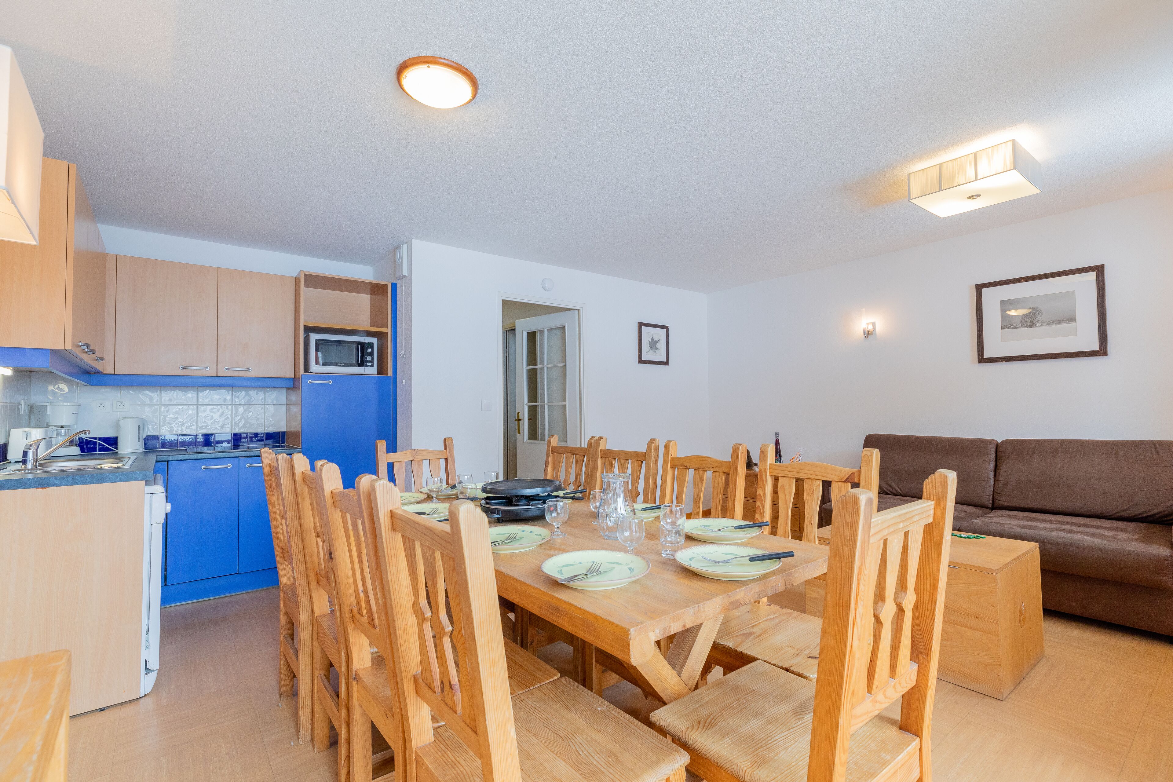 A modern kitchen with blue cabinets, a dining area with wooden chairs and a table set for six, and a brown leather sofa in the living area.