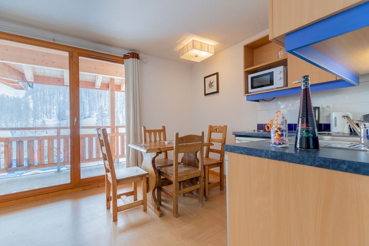 A wooden dining table with chairs, a kitchen area with a microwave, and a view of a snowy landscape through a sliding glass door.