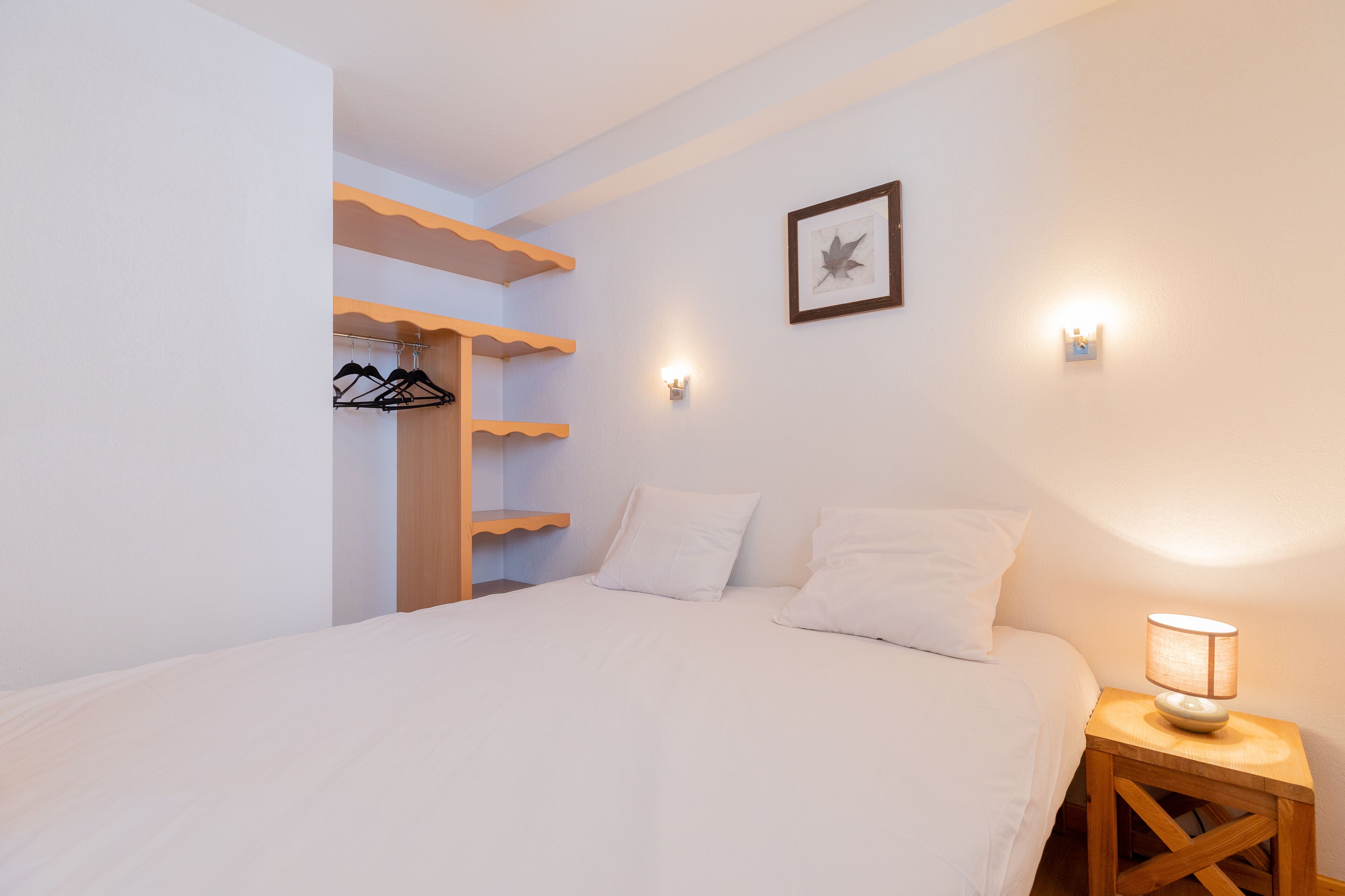 Un dormitorio con cama, mesita de noche con lámpara, estante en la pared con ropa y un cuadro enmarcado.